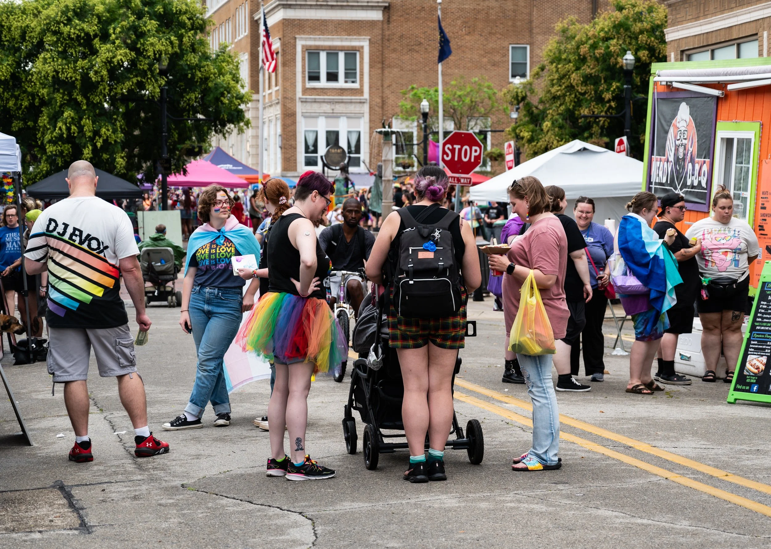 People gathered at an outdoor event (Anderson Pride), some wearing rainbow-colored clothing and accessories, with tents and stalls in the background, in an urban downtown street setting.