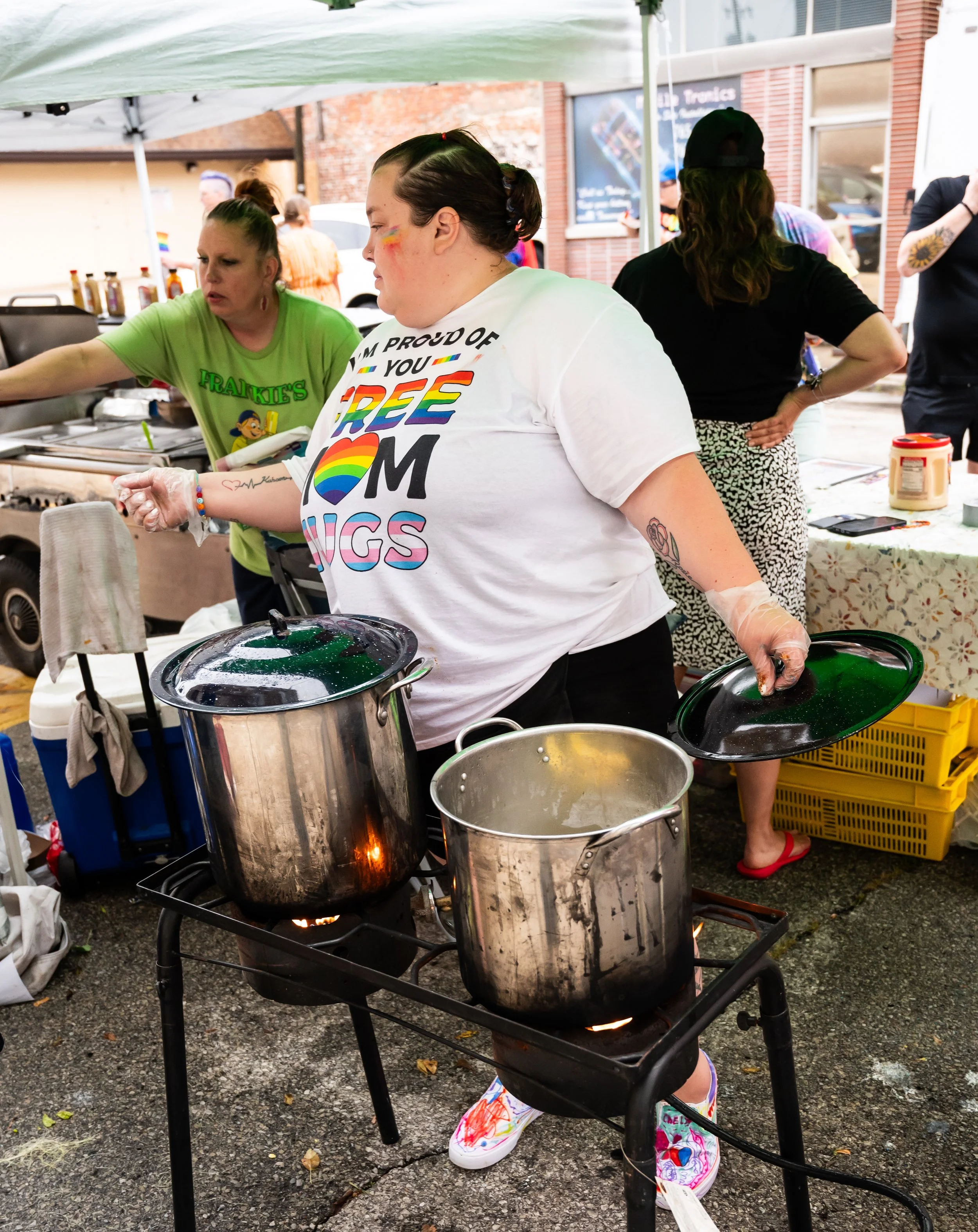Woman cooking at an outdoor event with two pots on a portable stove, wearing a white t-shirt with a rainbow-colored message, under a canopy, with people and food tables in the background.