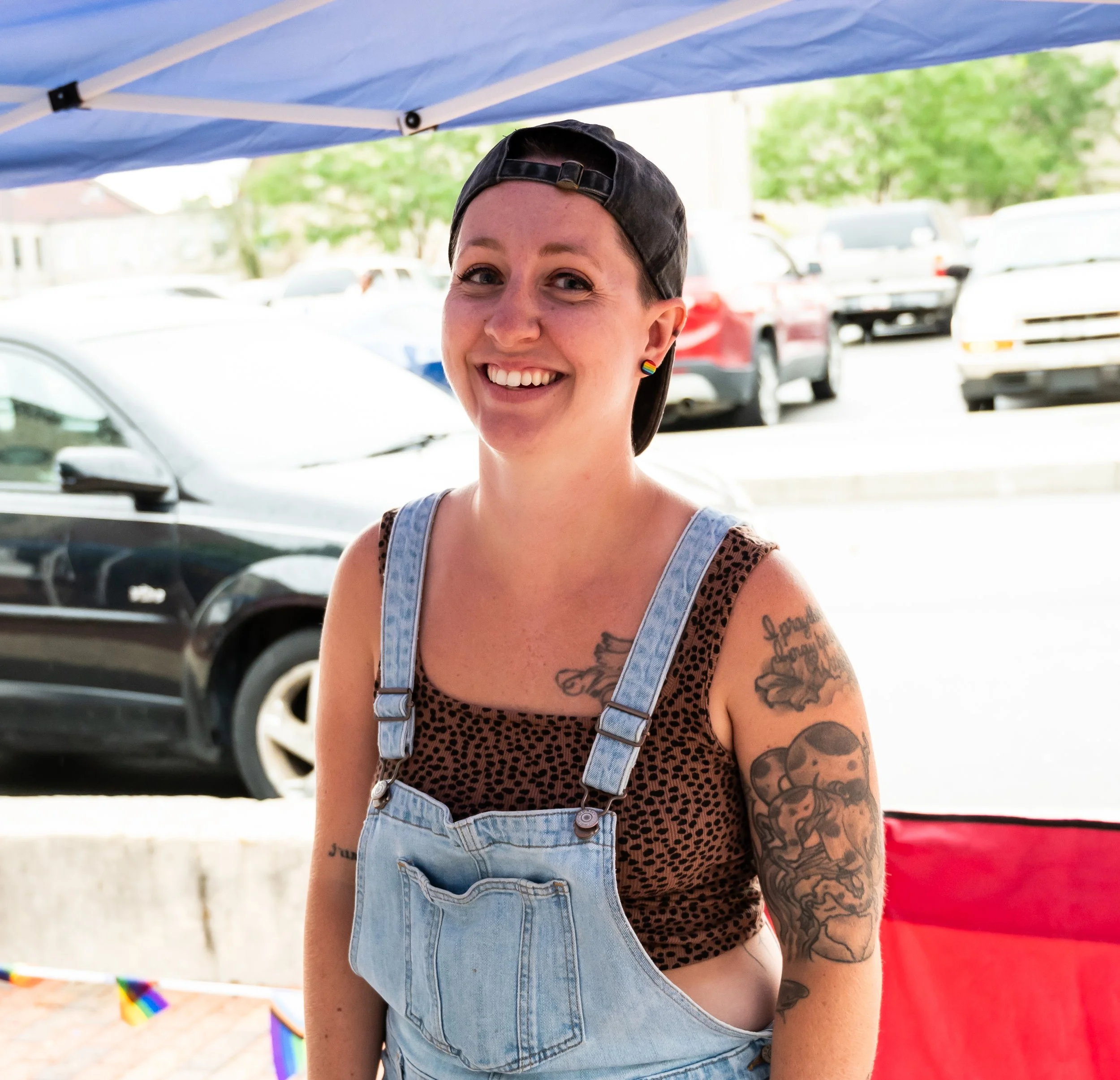 A smiling woman with short hair, wearing a black backward cap, rainbow earrings, a leopard print top, and light blue overalls, standing outdoors under a blue canopy with cars and greenery in the background.