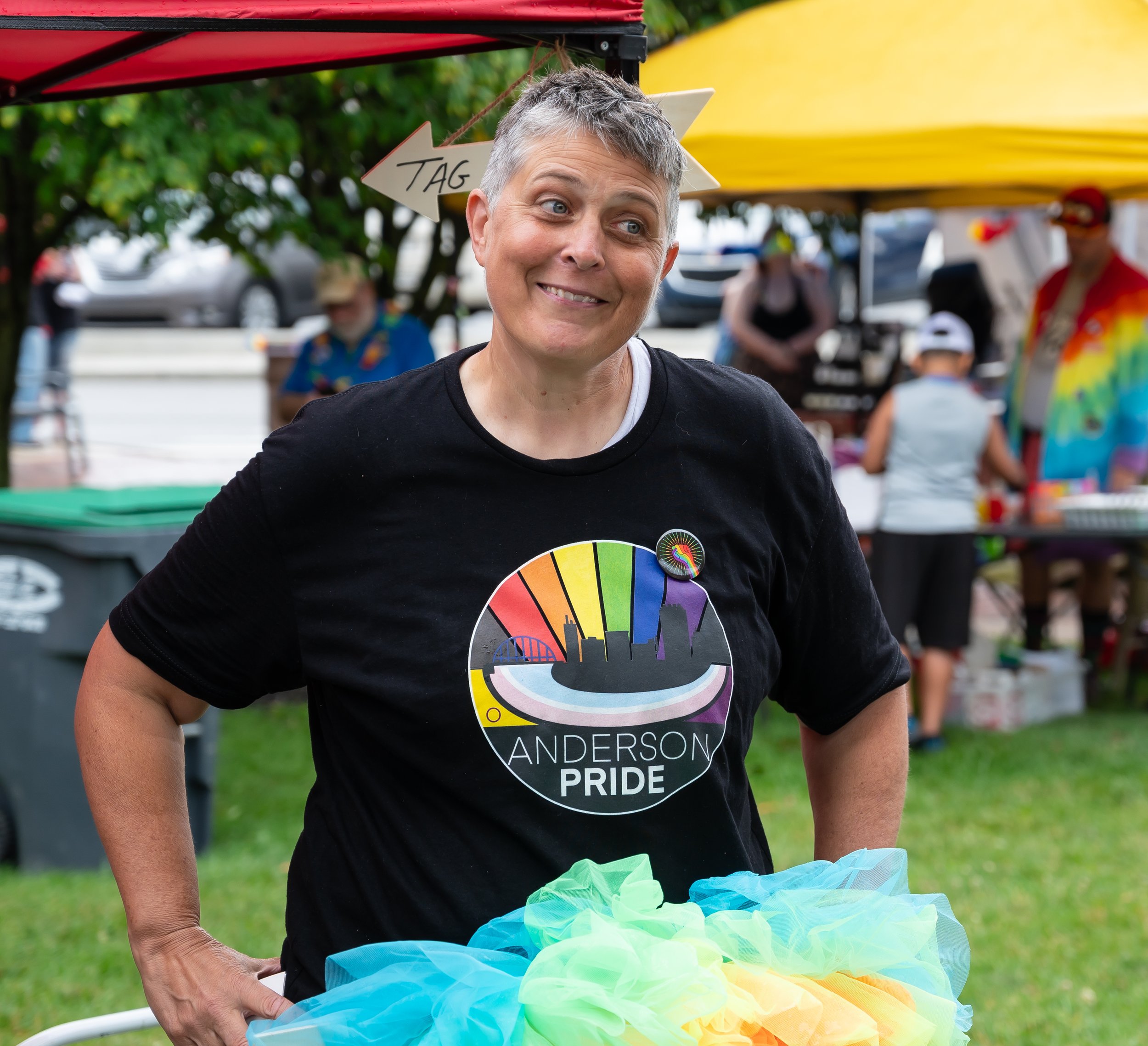 A person with short gray hair wearing a black shirt with a rainbow-colored cityscape and pride flag design, standing outdoors at a pride event with tents and other people in the background.