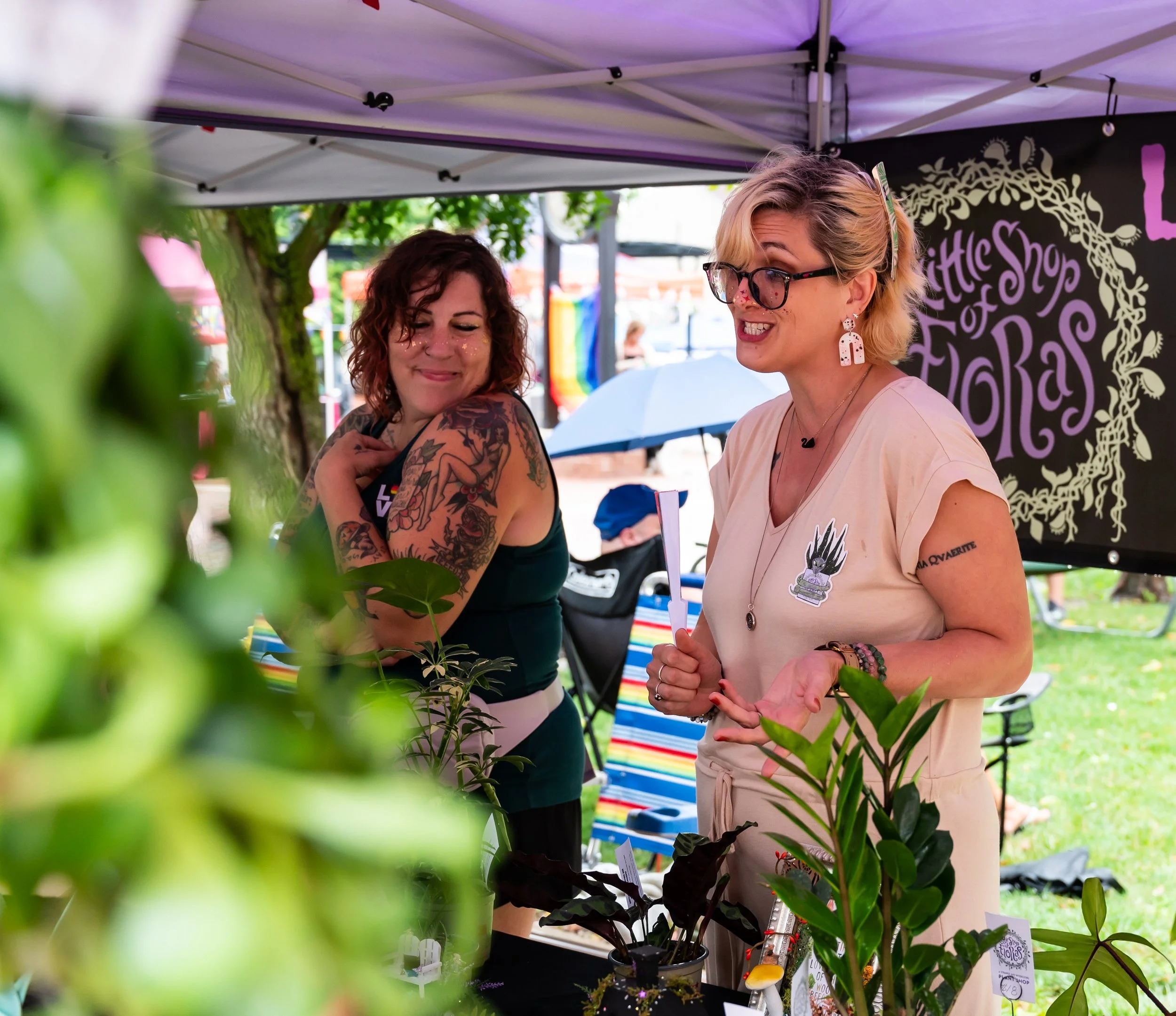 Two women engaging in conversation under a tent at an outdoor event, surrounded by green plants and a colorful striped chair, with a black banner behind them.
