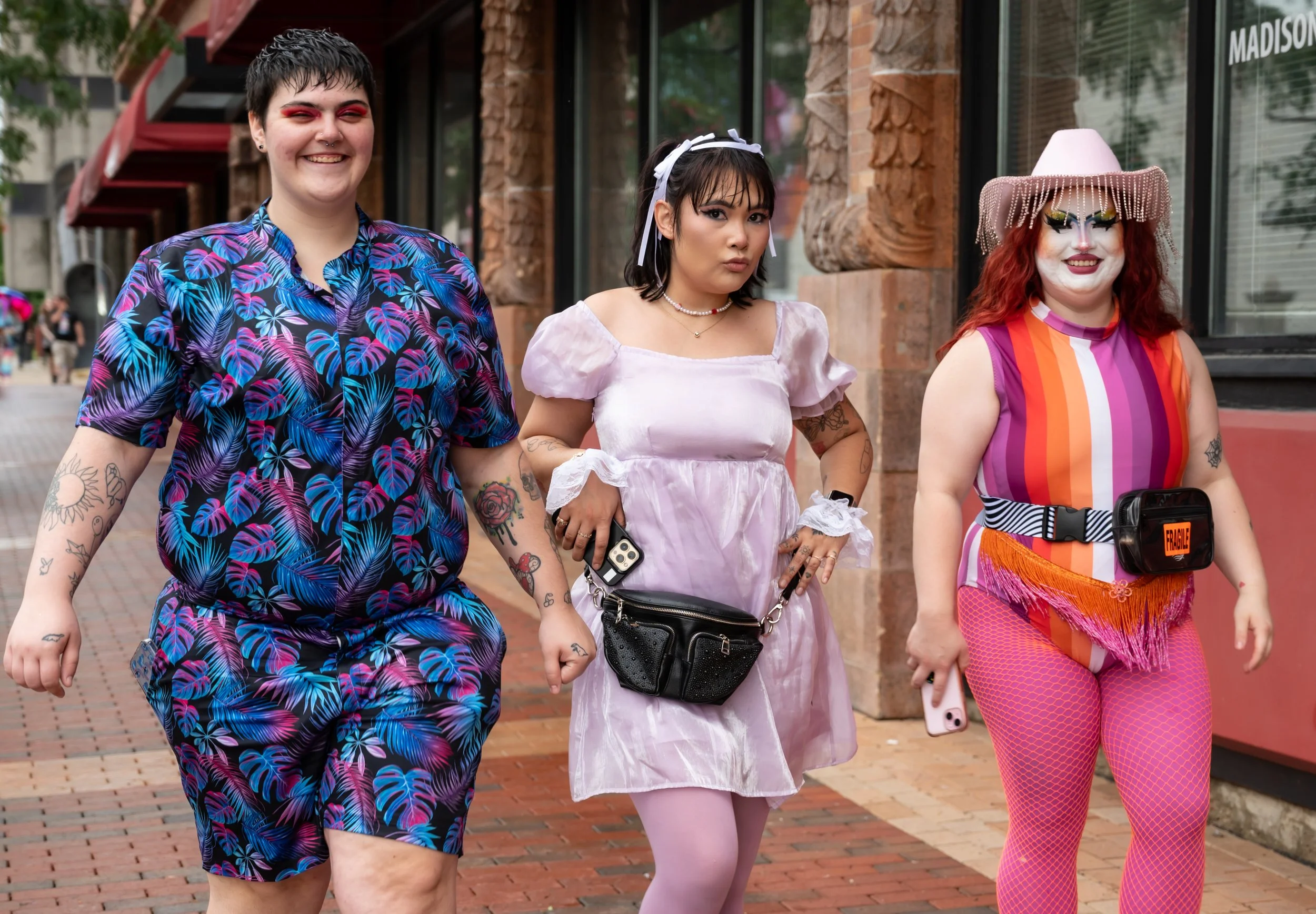 Three women walking outdoors, dressed in colorful and eclectic clothing, with tattoos visible on their arms, on a brick sidewalk in front of a restaurant or store with a large window.