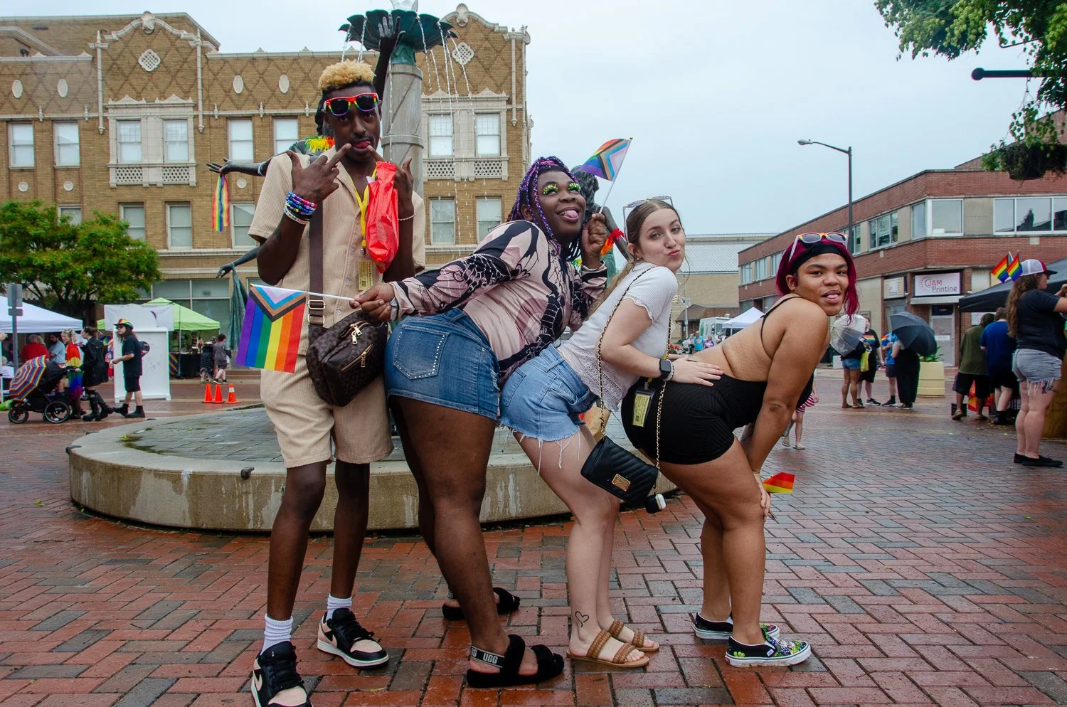Group of four diverse young adults posing playfully in a city square during a pride event, holding rainbow pride flags and wearing colorful accessories on a rainy day. In front of the "Three Graces".