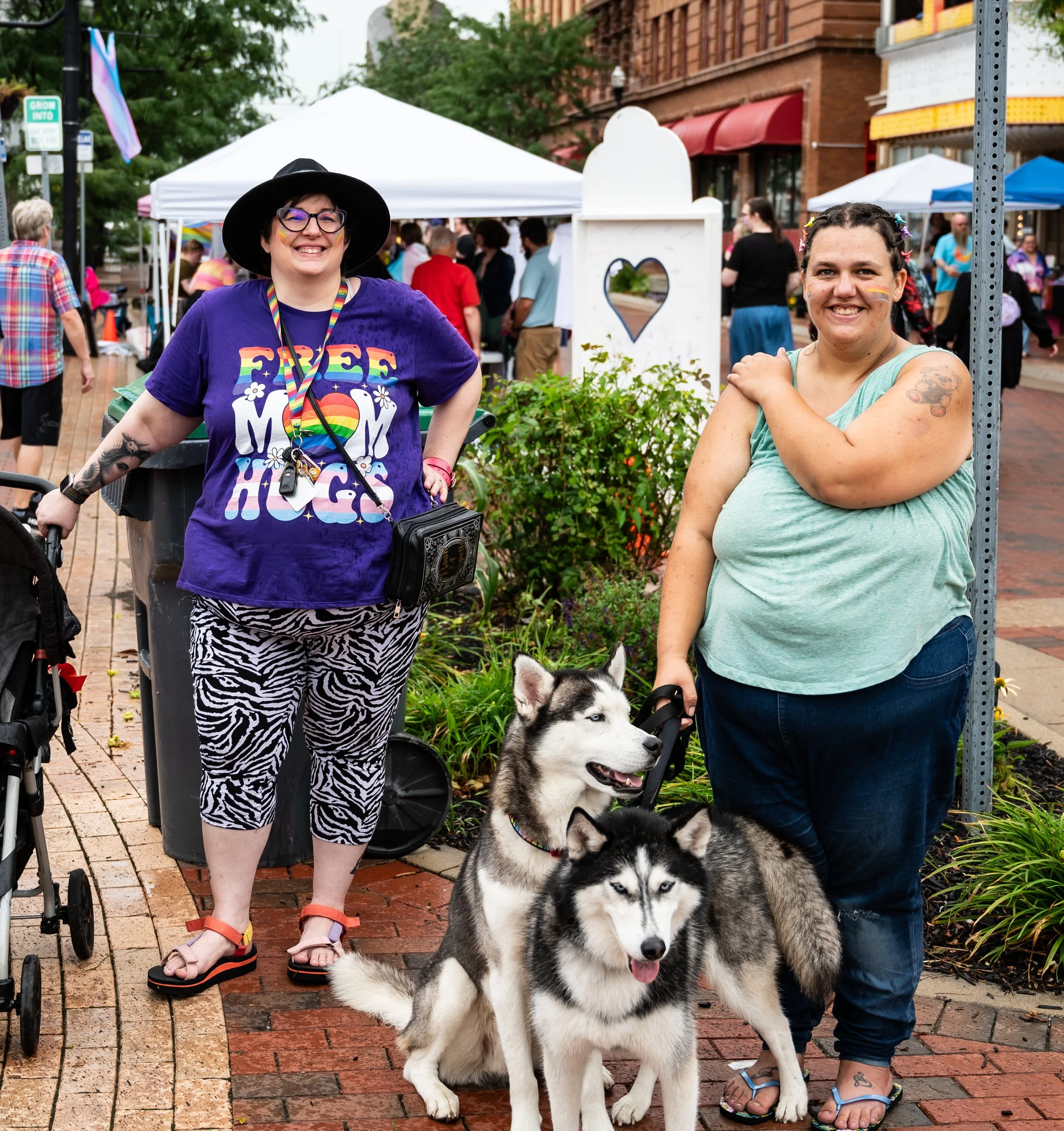 Two women smiling at Anderson Pride event with Huskies, outdoor street scene with tents and people in the background.