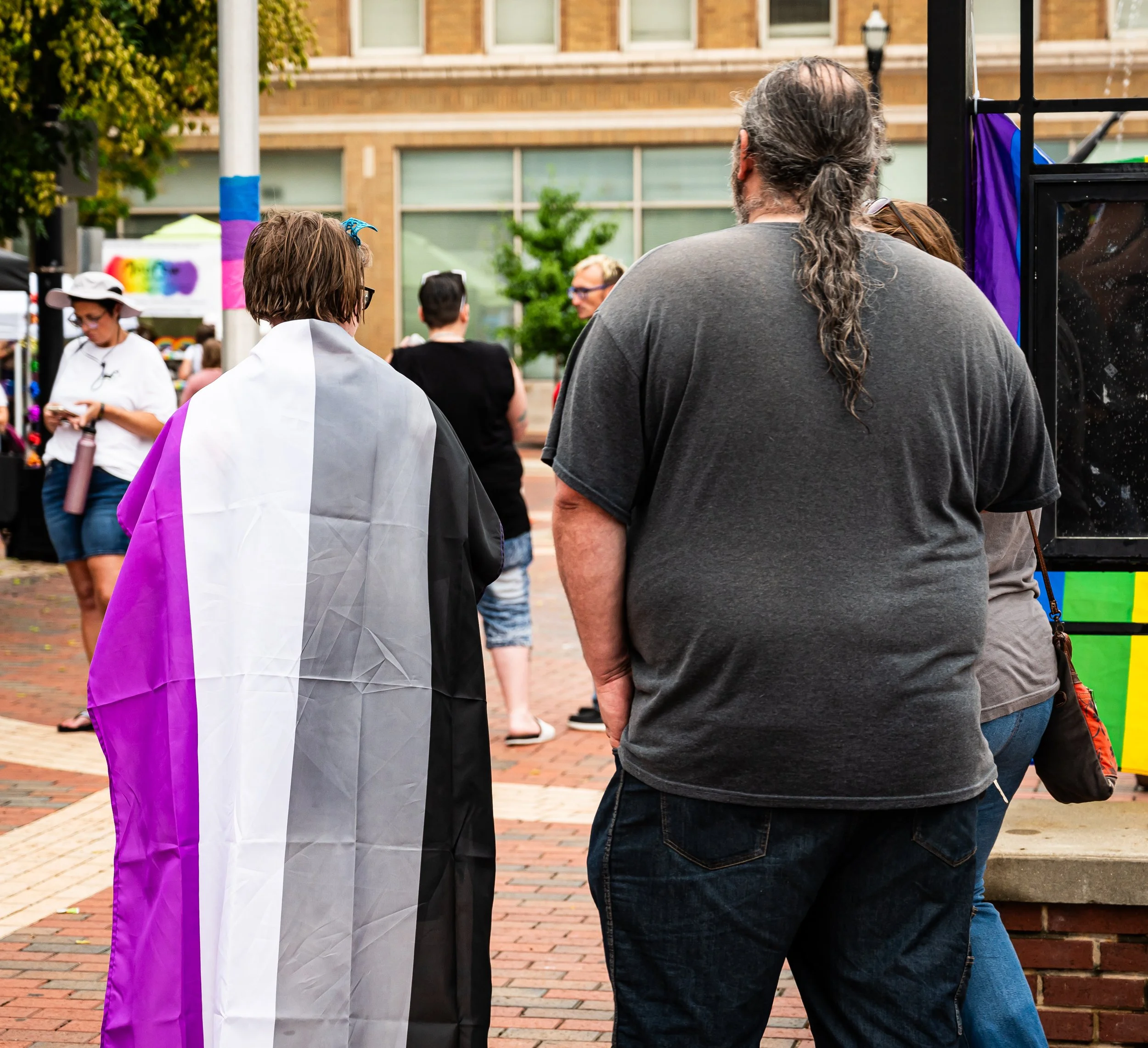 People gathered outdoors at an event, including a person with a rainbow pride flag draped over their shoulders.