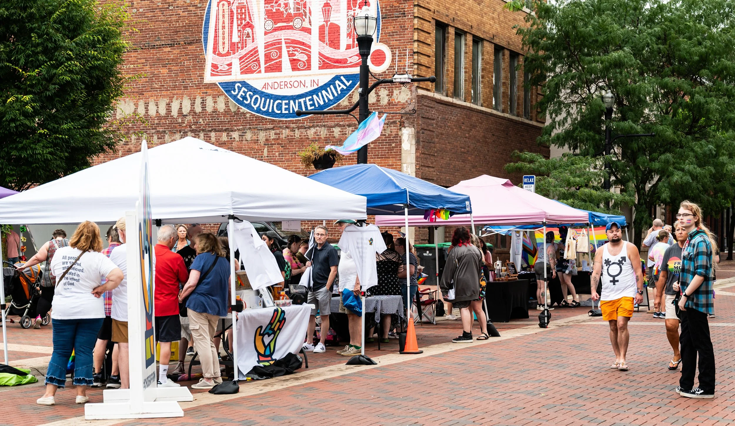 Outdoor street market with multiple booths and tents, with people browsing and shopping in front of a brick building and trees. Anderson Indiana, Anderson Pride. 