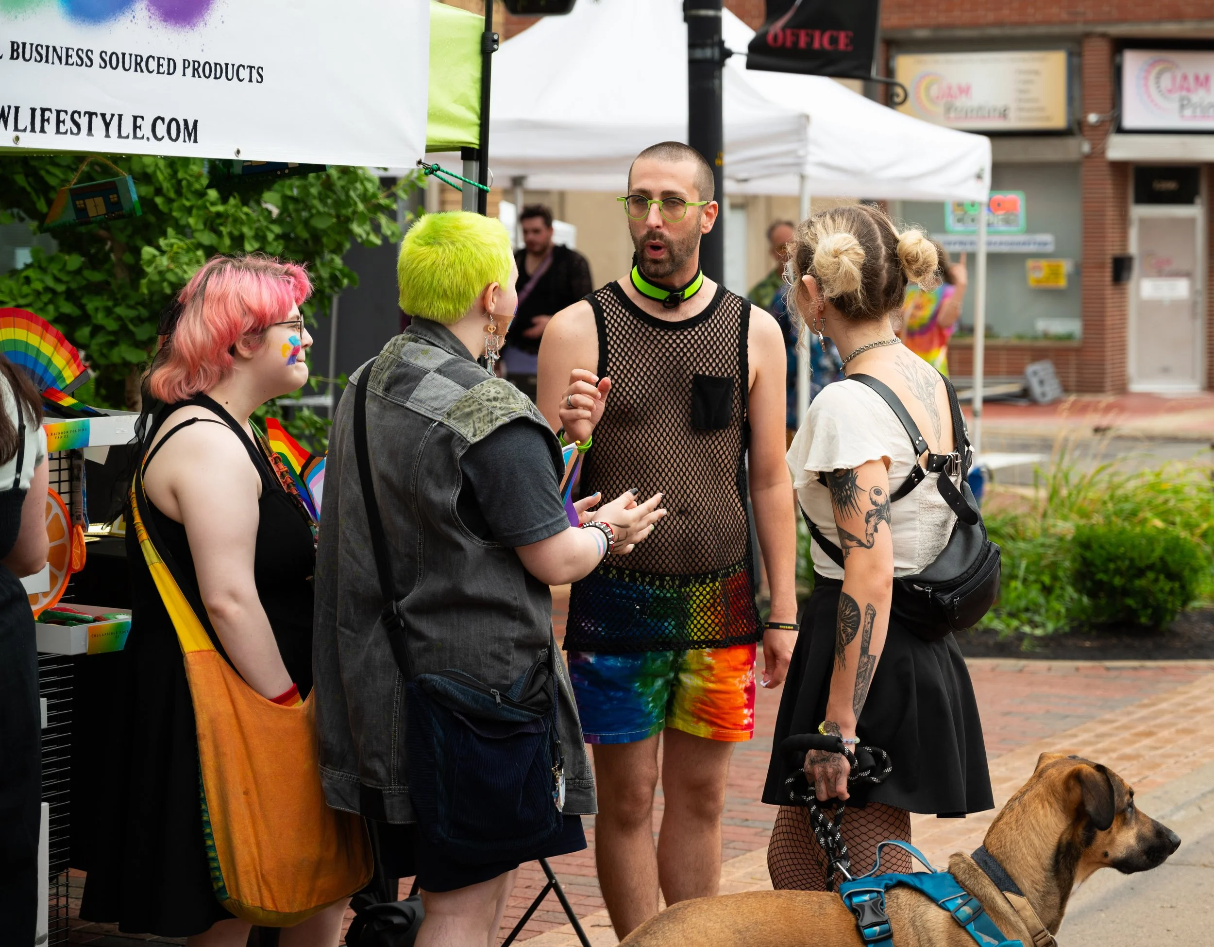 Group of five people with colorful hair and tattoos standing on a sidewalk at an outdoor festival, talking and holding a dog.