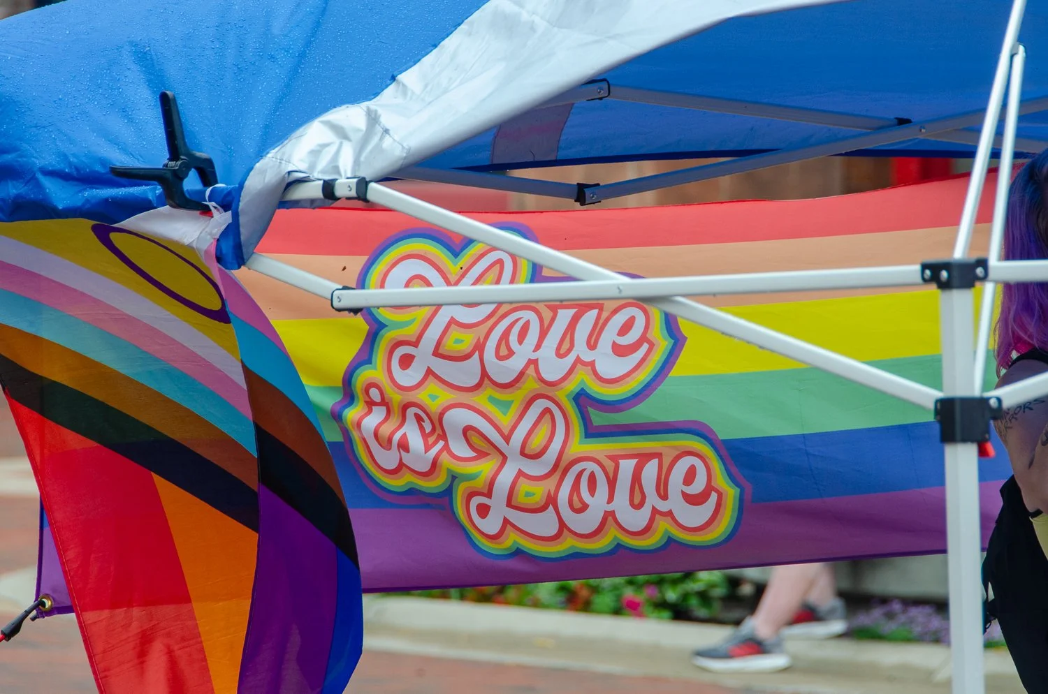 A colorful rainbow rainbow flag with the words 'Love is Love' written on it hanging under a blue canopy tent.