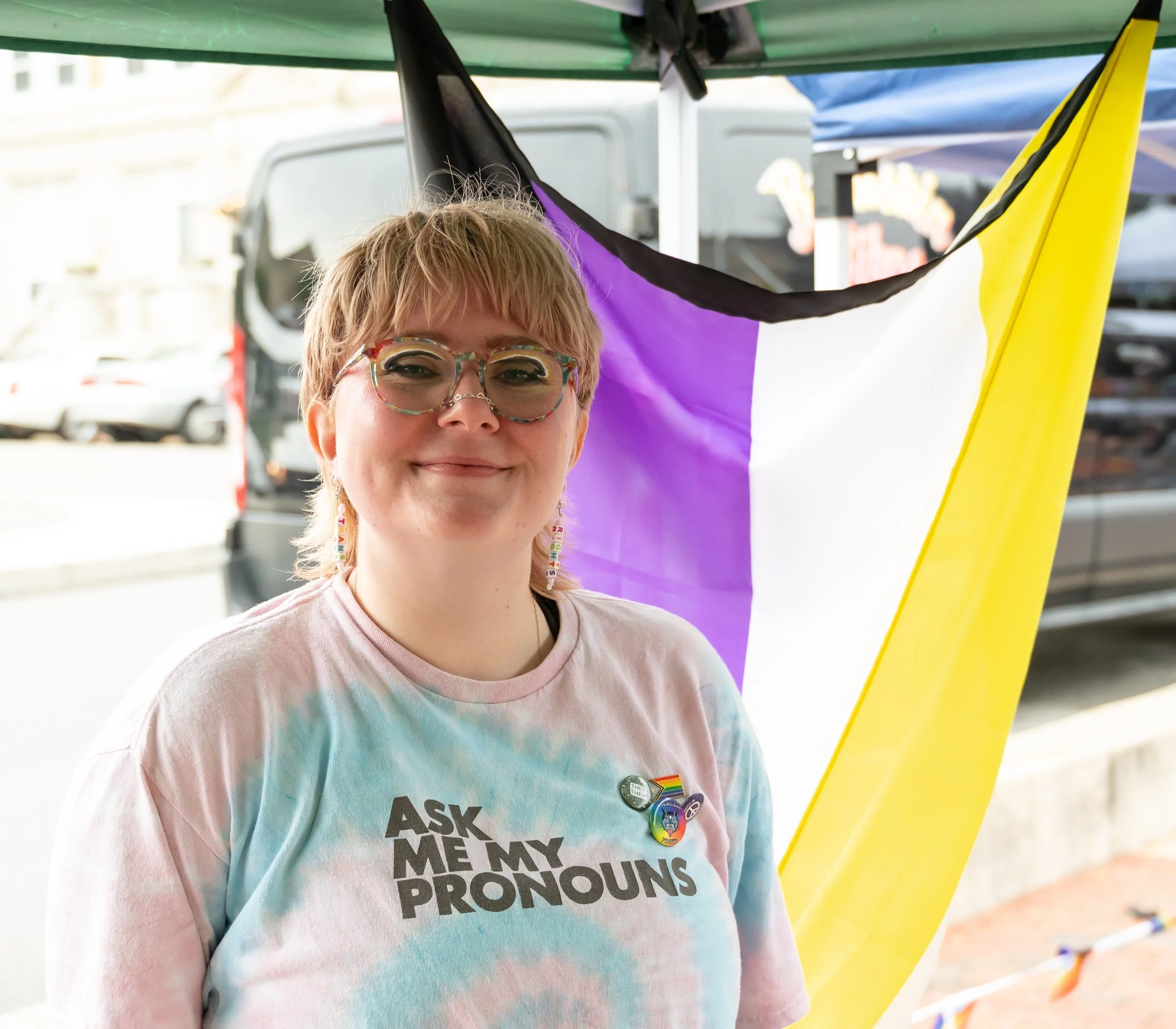 Person with short hair and glasses smiling, wearing a tie-dye shirt that says 'Ask Me My Pronouns,' standing in front of a rainbow flag at Anderson Pride.