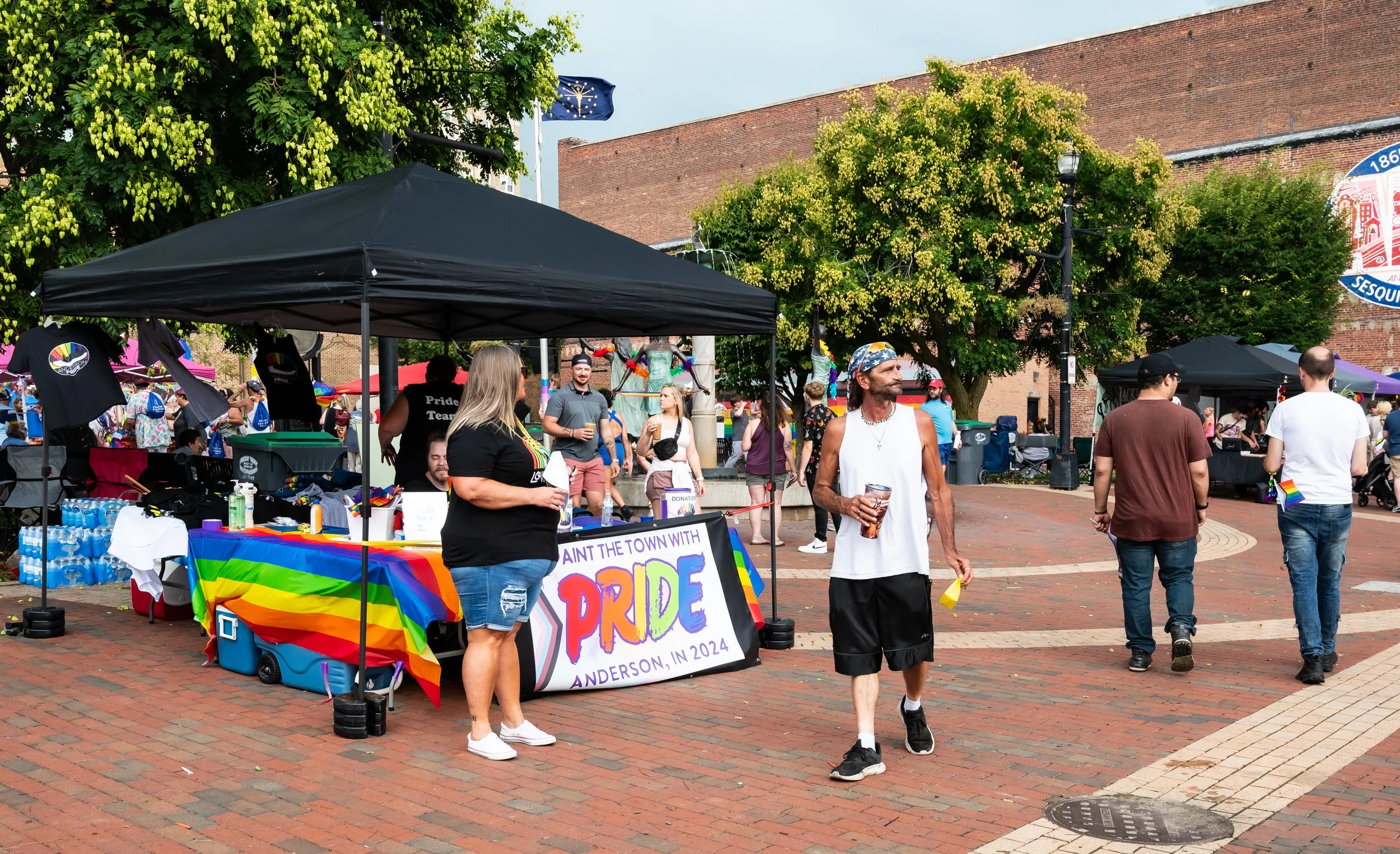 Anderson Pride booth decorated with rainbow flags, and a sign that reads 'Paint the town with PRIDE Anderson, IN 2024.' Visitors walk around this outdoor market, some carrying beverages, with trees and a brick building in the background.