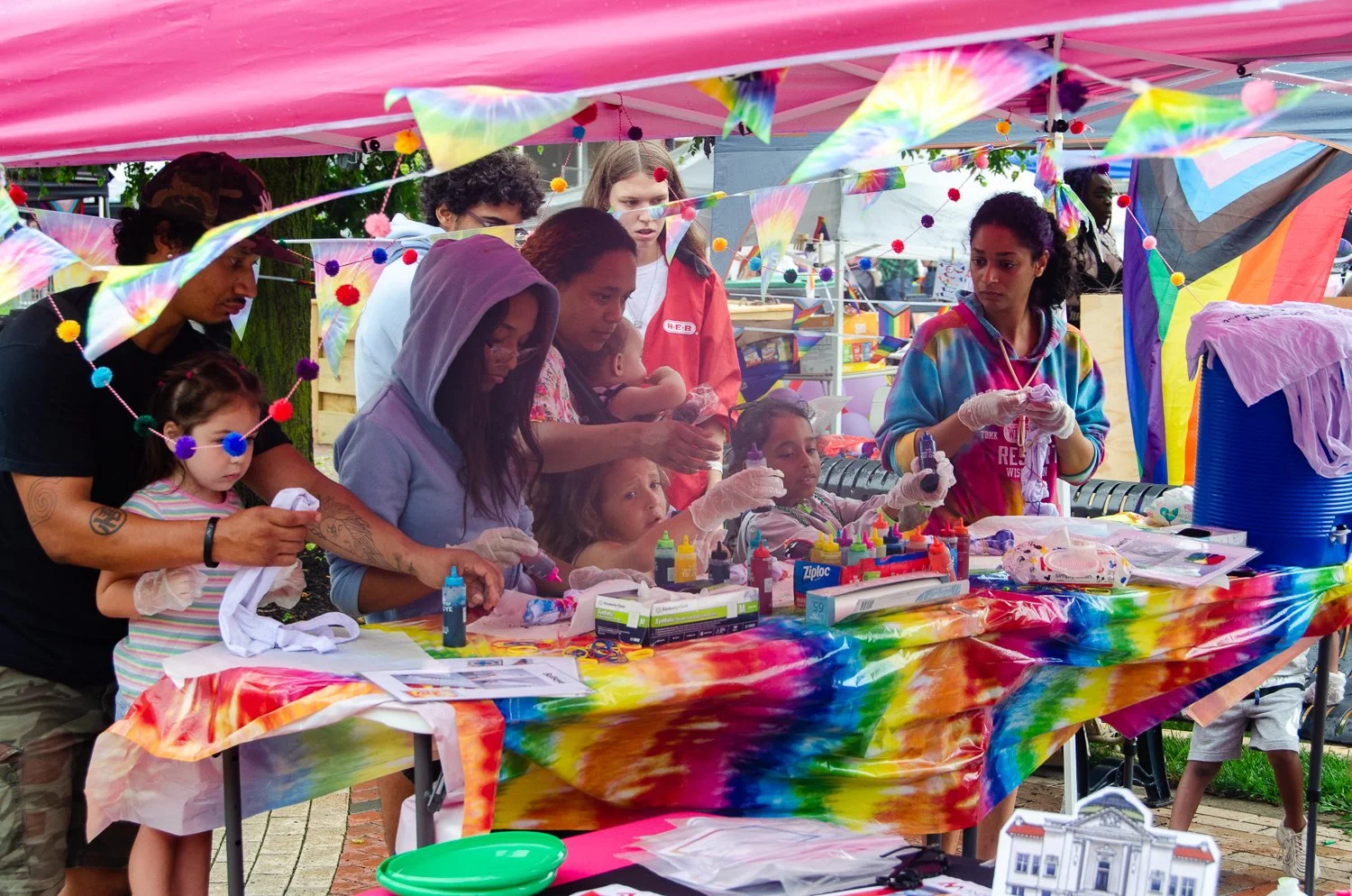 People of various ages and ethnicities gathered under a colorful tie-dye canopy at a communal outdoor arts and crafts event, working on various craft projects with supplies on a rainbow-colored table.