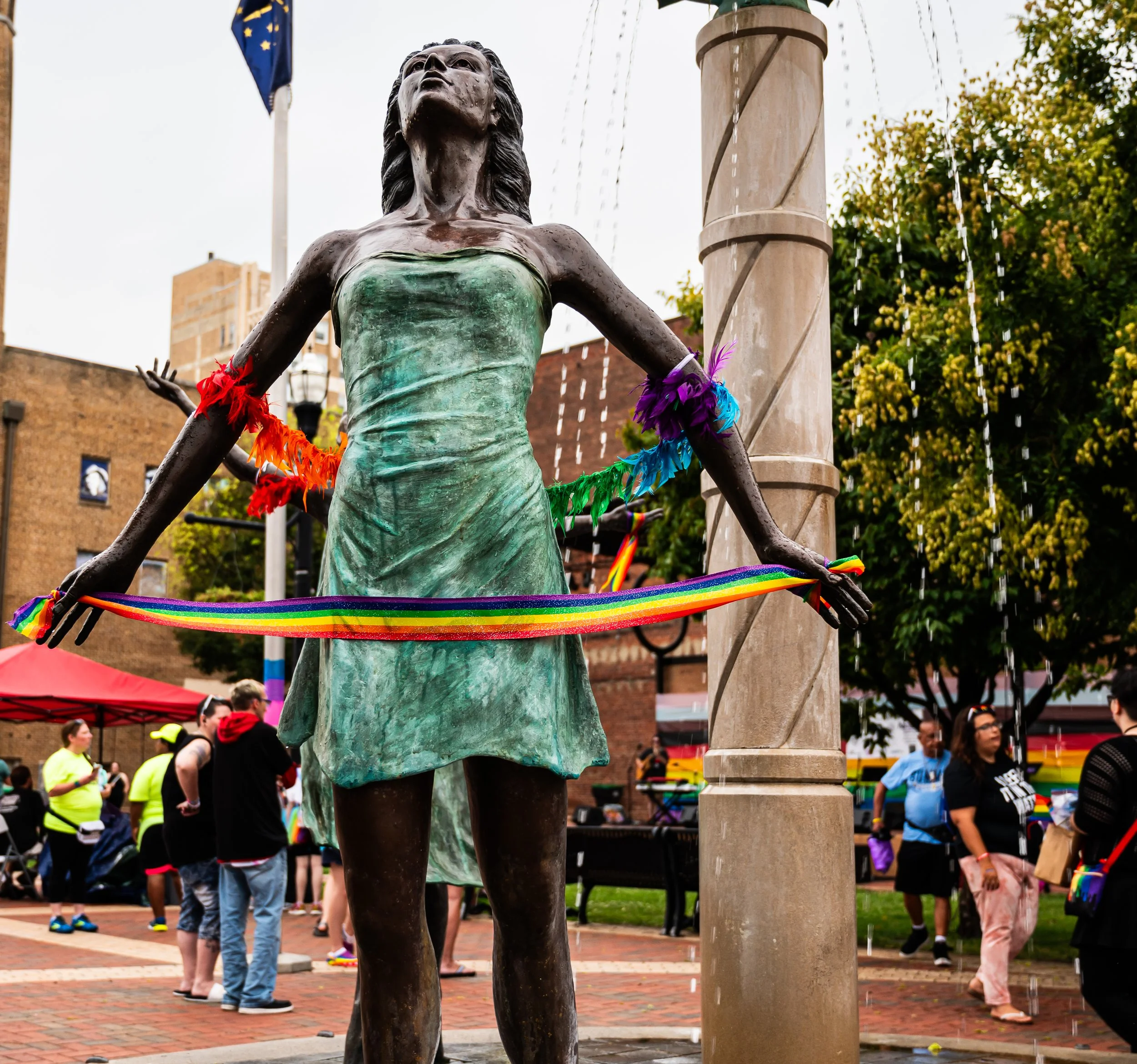 Statue of a woman holding a rainbow rainbow-colored ribbon at a pride event, decorated with rainbow-colored feathers and beads, with people gathered in the background. "Three Graces" Anderson Indiana. Dickmann Town Center. 