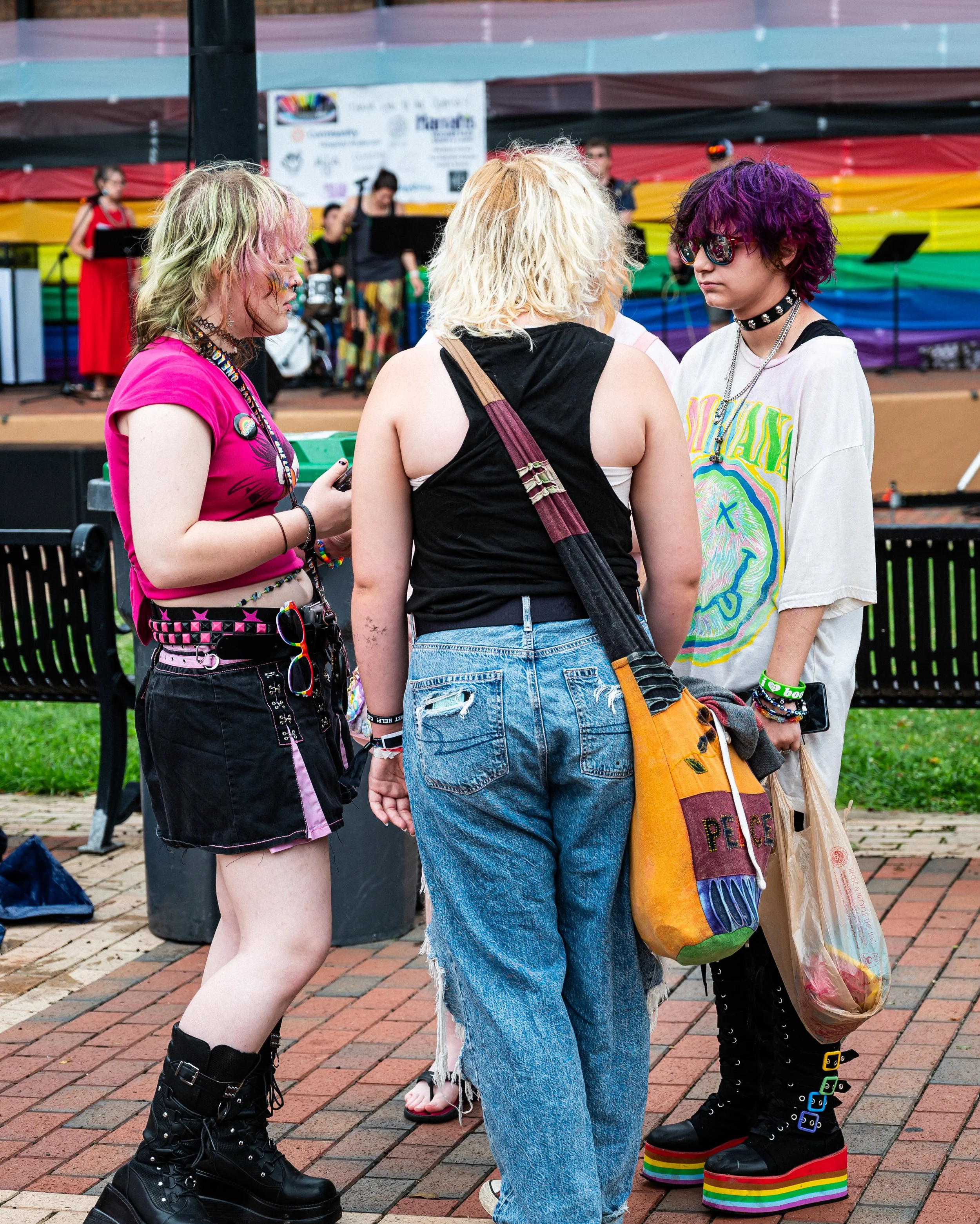 Three young women with colorful, alternative styles standing and talking outdoors at a pride event, with a stage and rainbow decorations in the background.
