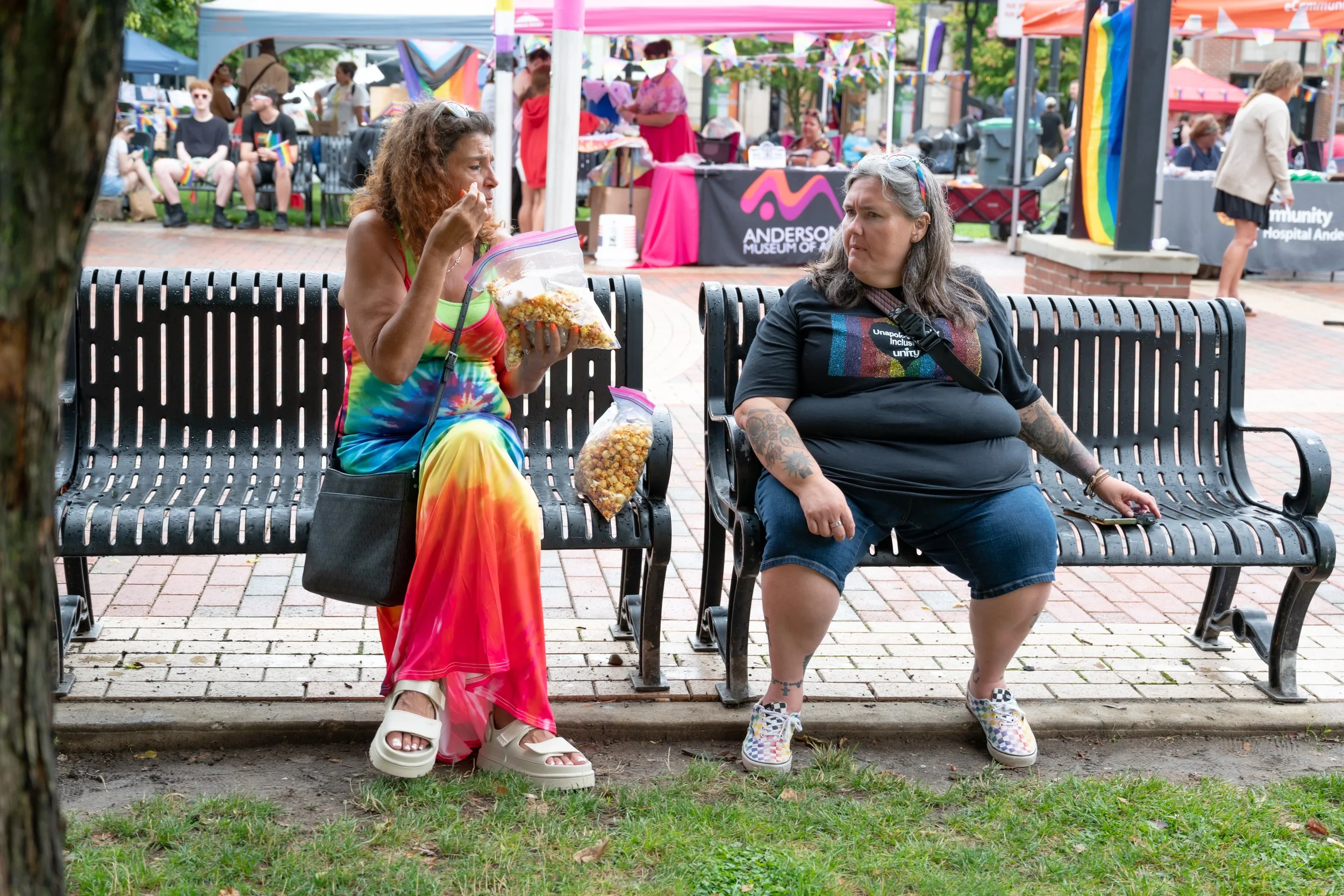 Two women sit on park benches at an outdoor community event with colorful tents and booths in the background. One woman, wearing a rainbow tie-dye dress, is eating popcorn from a clear plastic bag, while the other woman, dressed in a black shirt with
