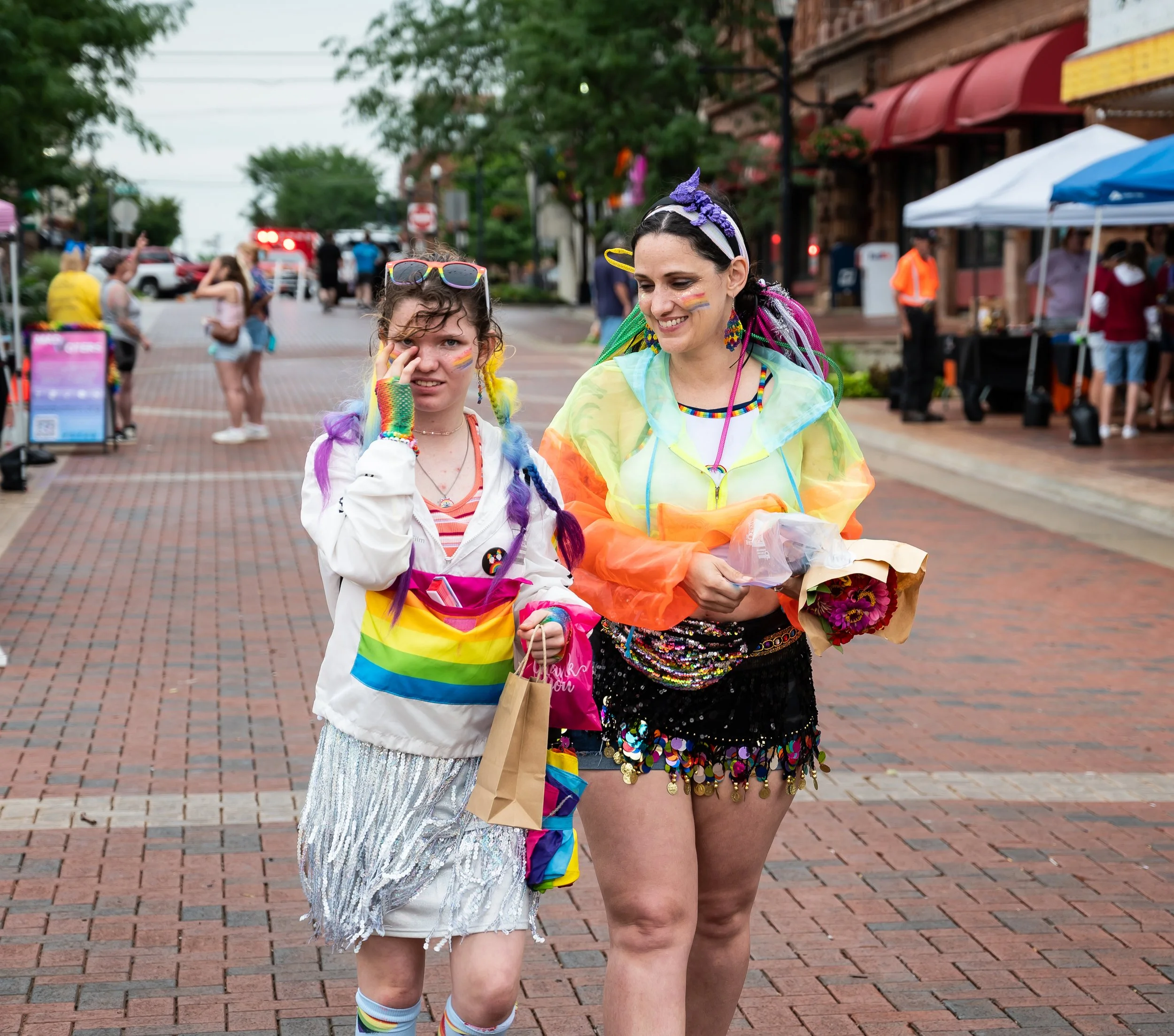 Two women walking together during Anderson Pride event, both decorated with rainbow accessories and face paint, one with rainbow braids and the other with rainbow stripes on her face, carrying flowers and shopping bags.