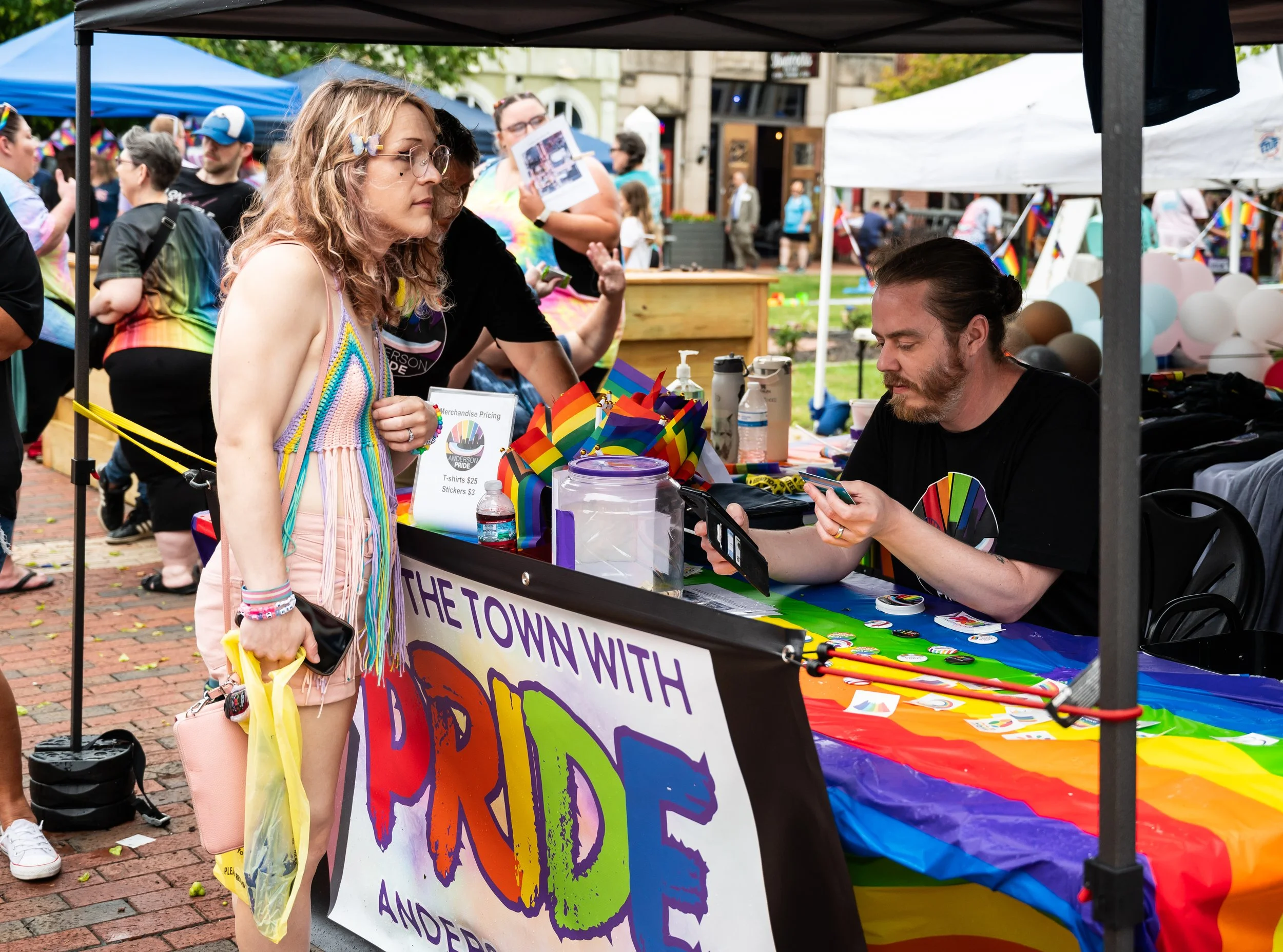 A woman in colorful, rainbow-themed clothing is purchasing items at a pride event booth. The booth has rainbow decorations, pins, and informational signs, with a sign reading "The Town with Pride". Other attendees are visible in the background, histo