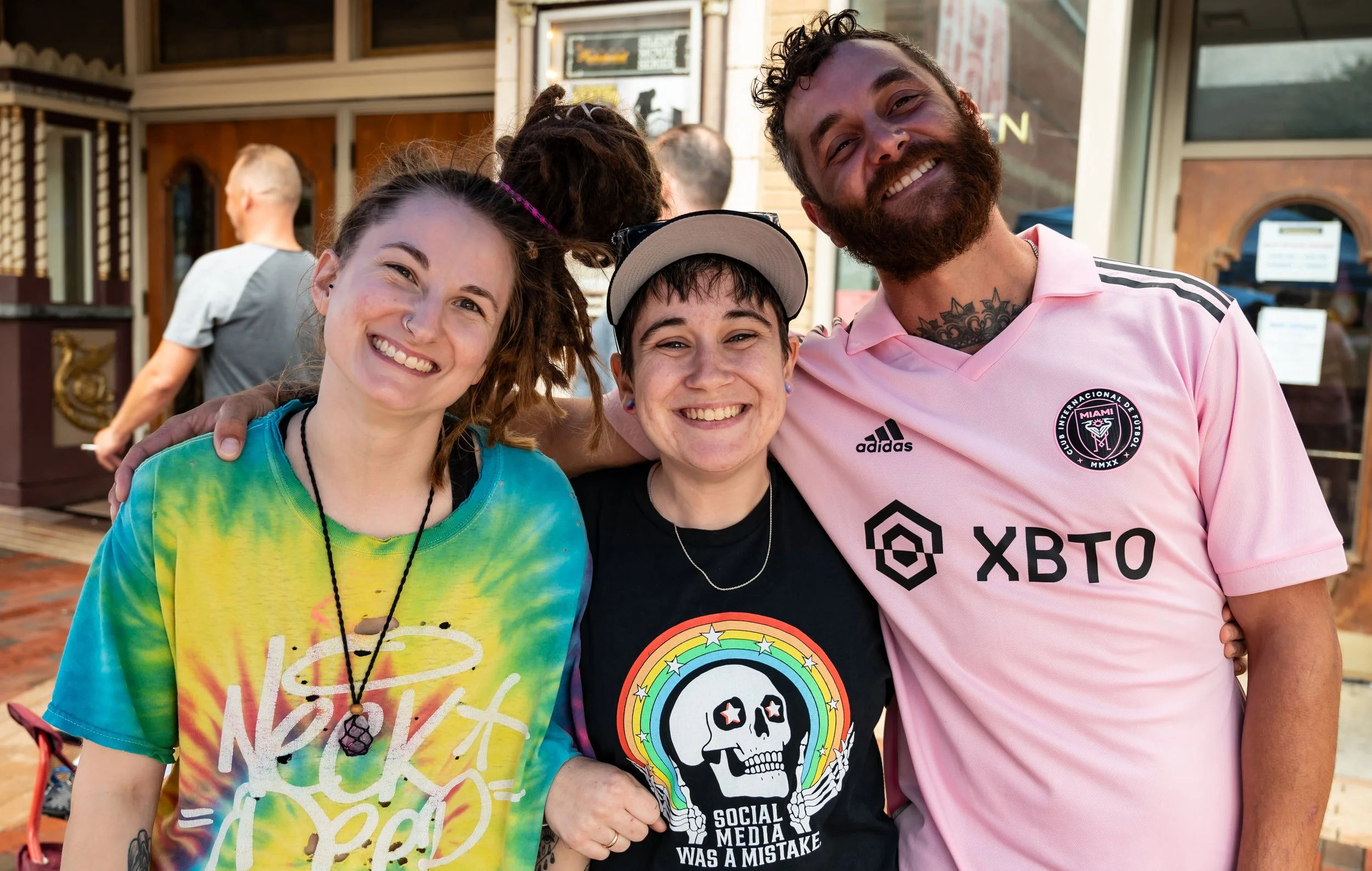 Three people smiling and posing closely together outdoors, with a man on the right wearing a pink soccer jersey and two women in casual clothing on the left, one with dreadlocks and the other wearing a cap.