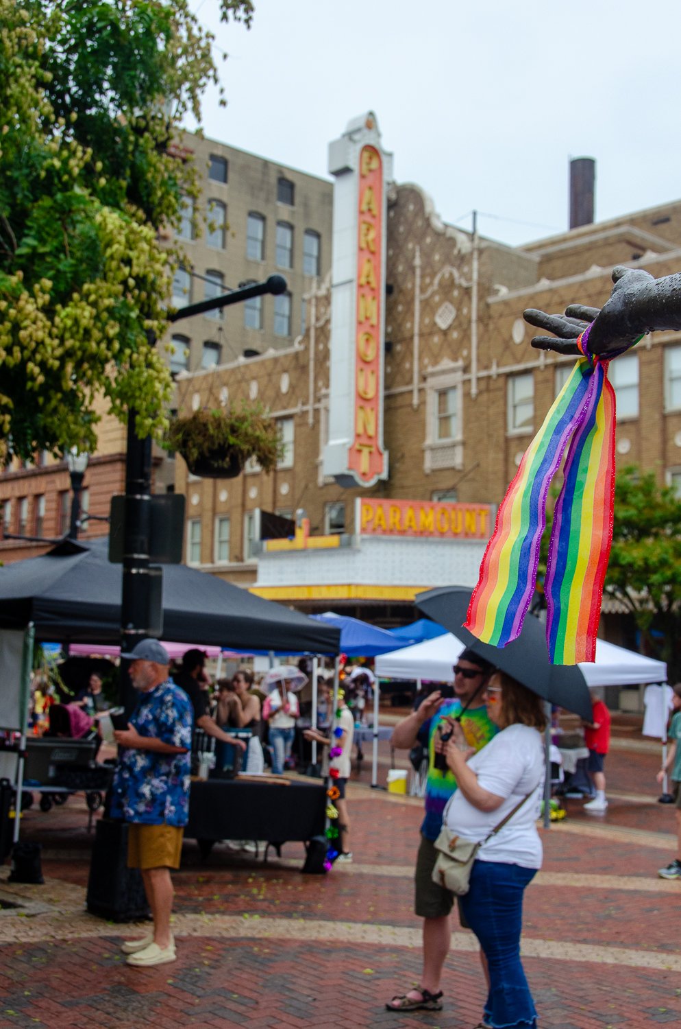 Street scene outside the Paramount Theater in Anderson Indiana with people shopping at outdoor stalls, with one person holding an umbrella and rainbow ribbons attached to a statue's hand, and a sign with the theater's name in the background.