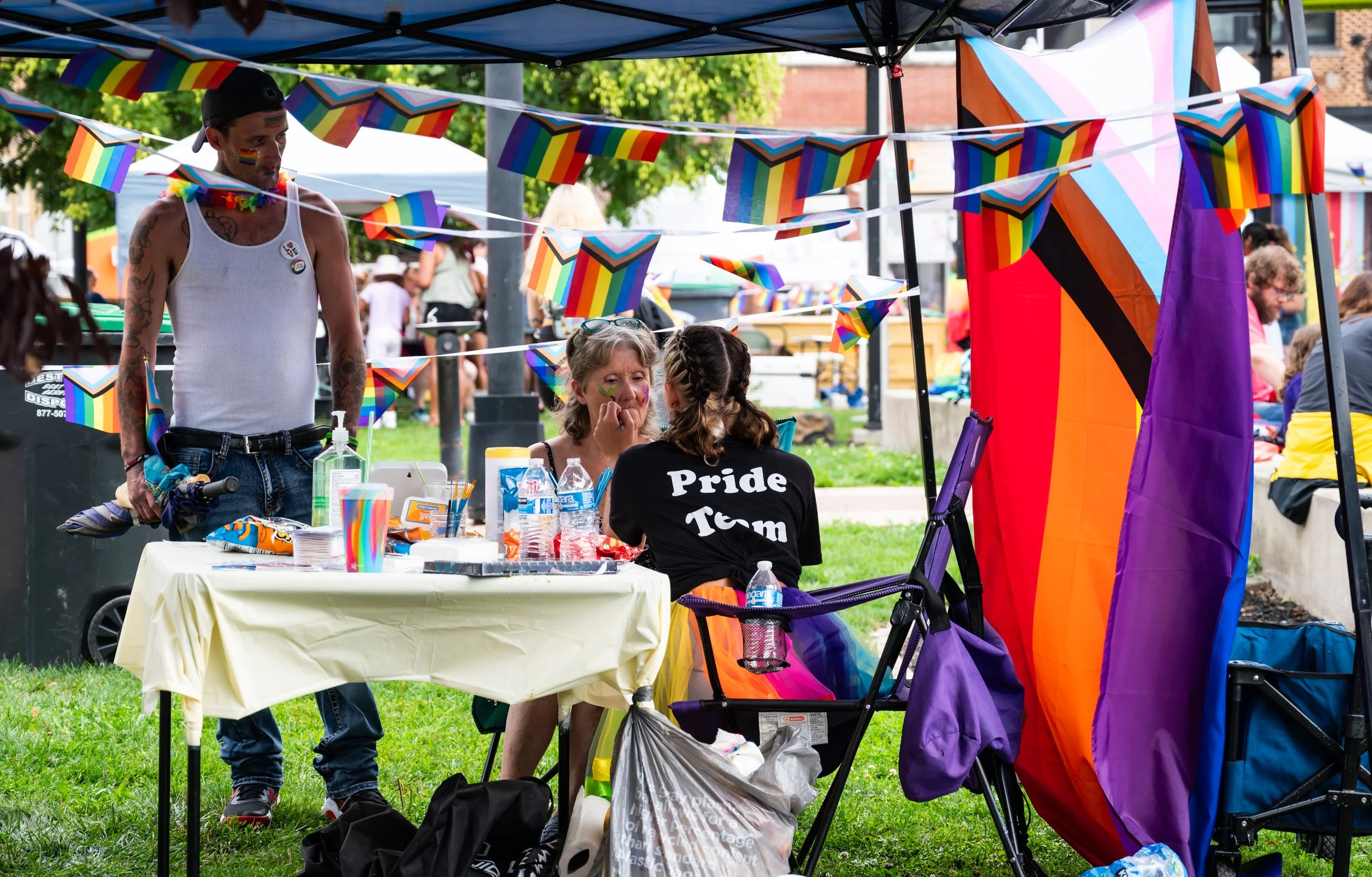 People at a Pride celebration under a tent decorated with rainbow flags, with a table displaying resources, water bottles, and snacks, and a rainbow pride flag draped on the side of the tent.