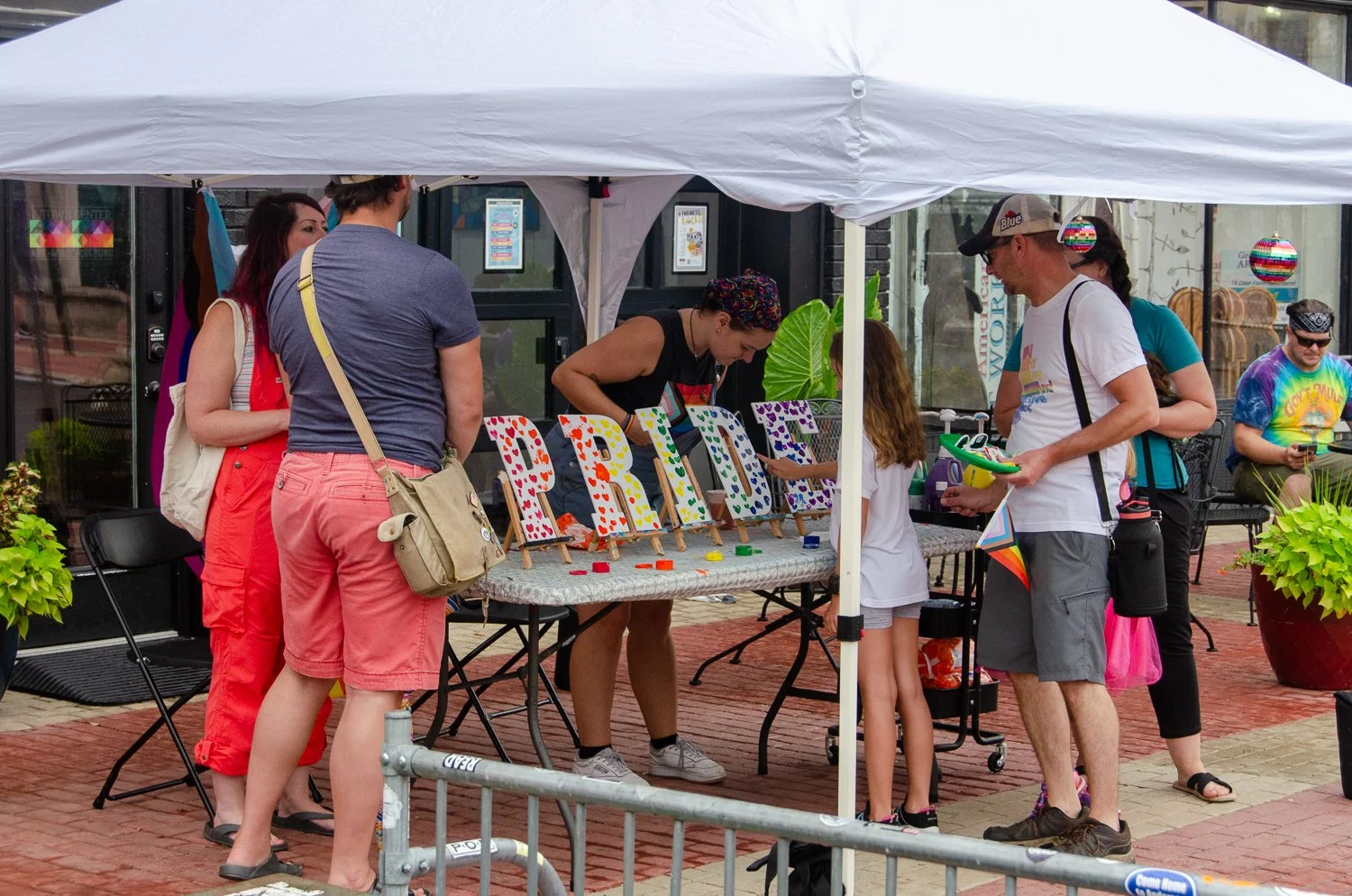 People gathered around a booth at an outdoor event, where they are painting decorative signs that spell out 'PRIDE'.