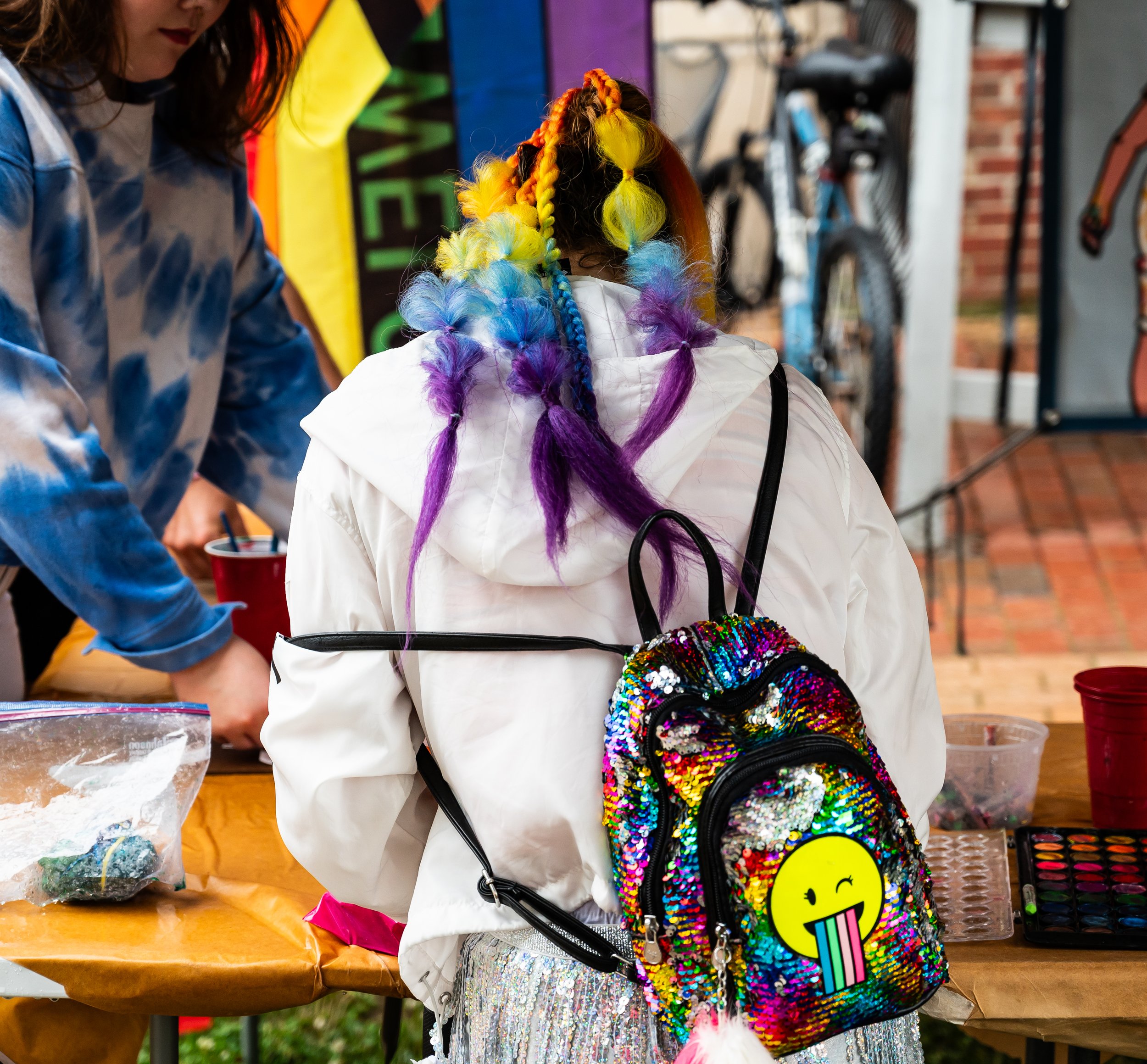 Person with colorful rainbow hair, a white jacket, and a sequined backpack with a yellow smiley face, standing at a table with art supplies.