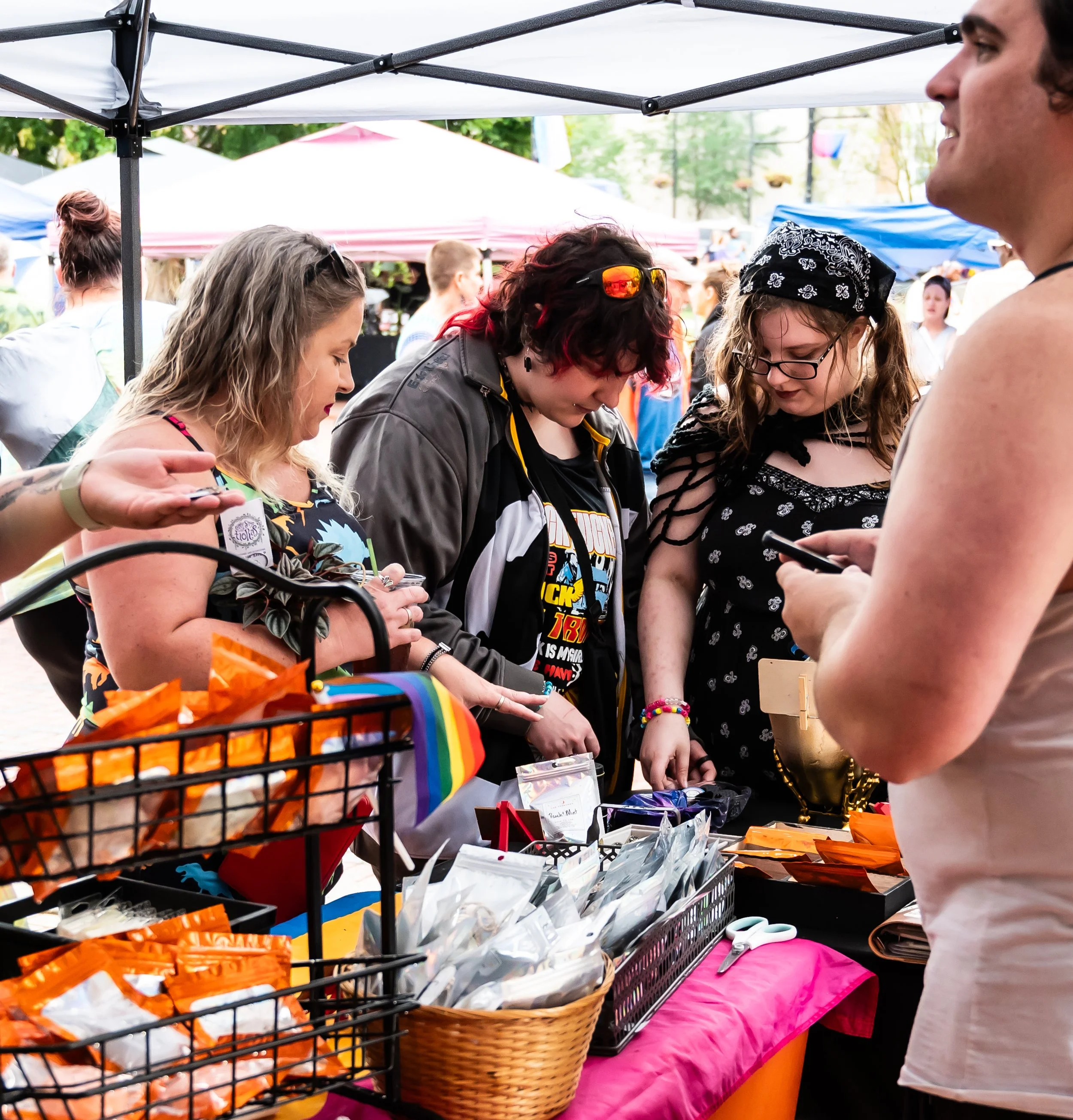Group of people at an outdoor marketplace or craft fair, browsing items on a table under a canopy, with several colorful tents in the background.
