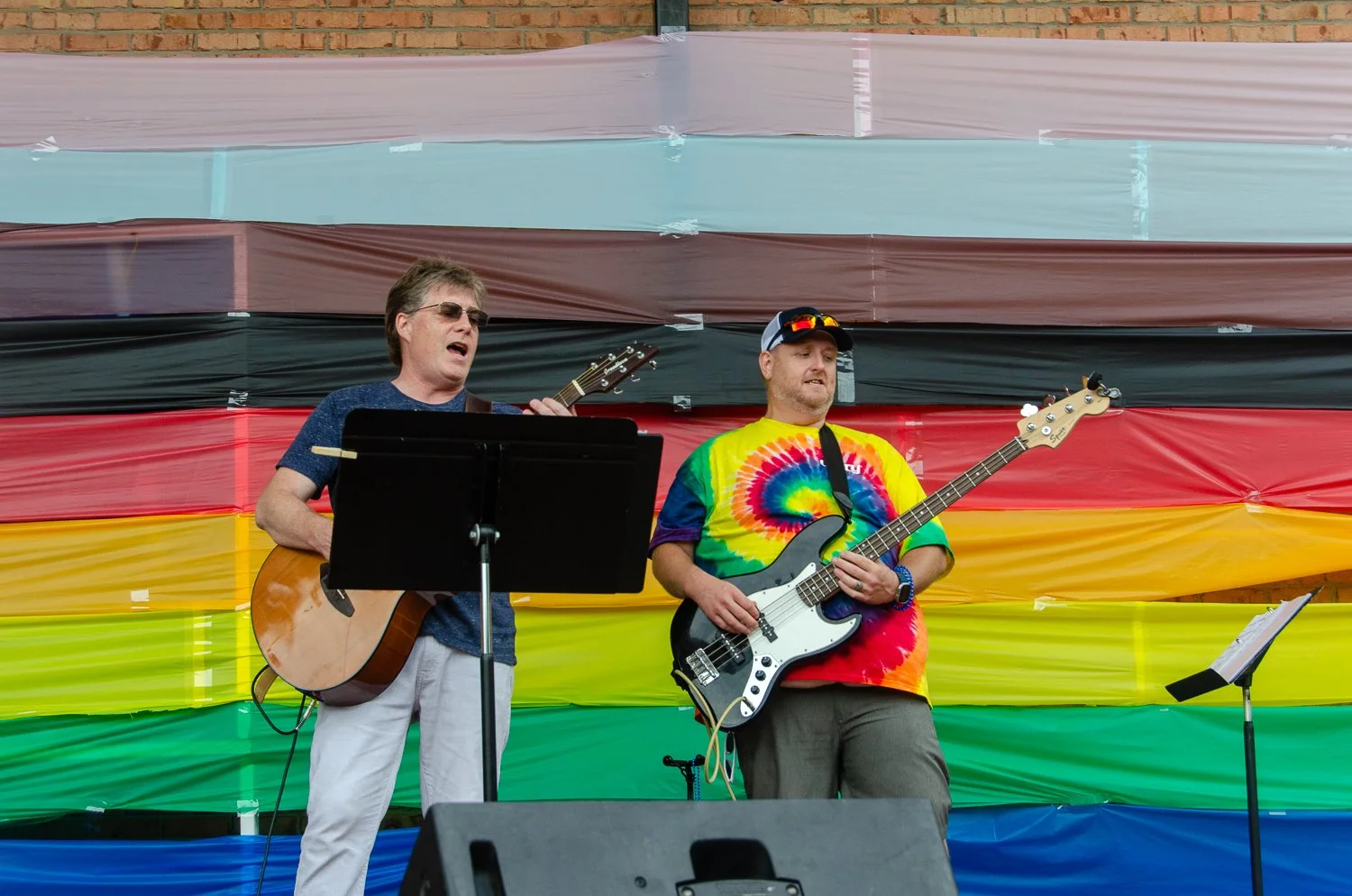 Two musicians performing on stage in front of a large rainbow-colored striped backdrop, one playing an acoustic guitar and the other playing an electric bass, both singing and wearing casual clothing.