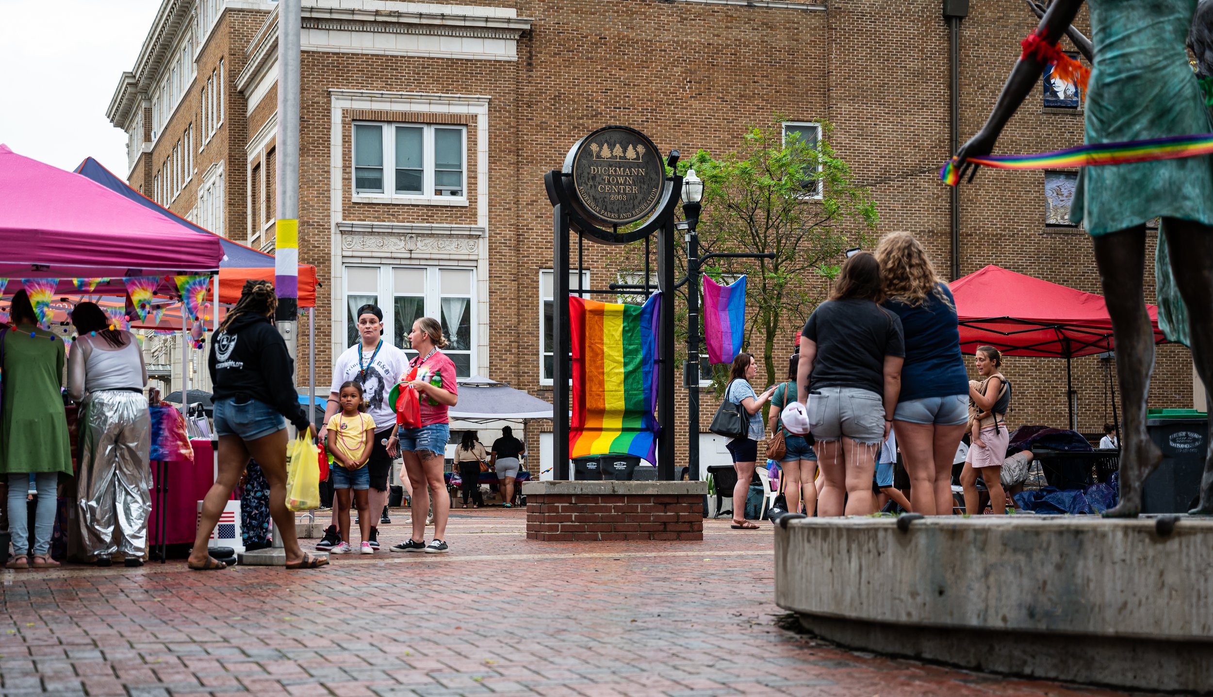 People gathered at Anderson Pride, in Anderson Indiana in Dickmann Town Center with tents, rainbow flags, and statues, during daytime.
