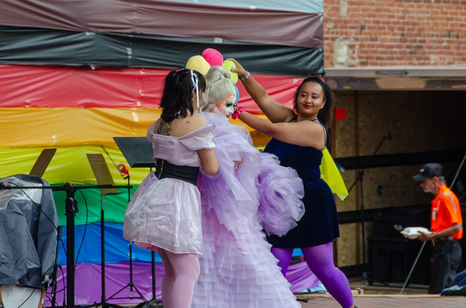Two women, one dressed as a fairy godmother and another as a fairy, assist a person in a large, purple, layered dress during an outdoor event on a stage with rainbow-colored decorations.