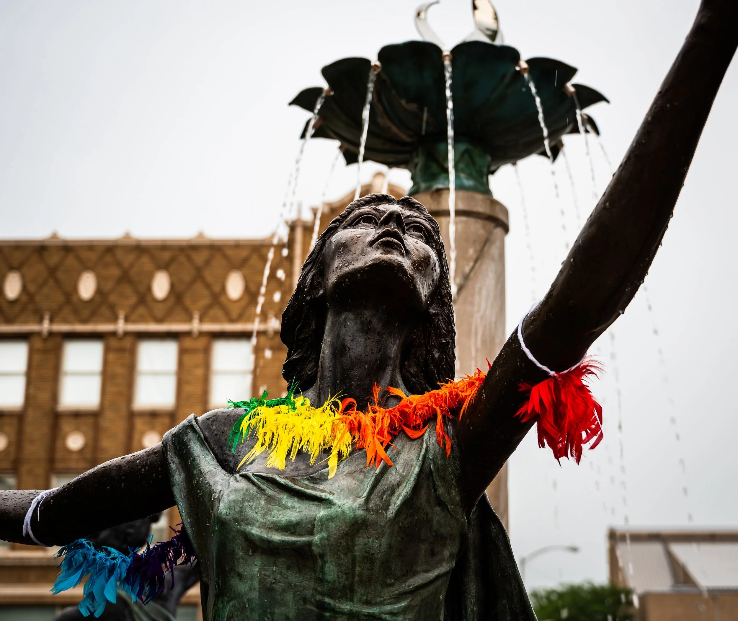 Statue of a "Three Graces" with rainbow-colored feathers around her neck in front of a fountain and brick building, Dickmann Town Center, in Anderson Indiana. 