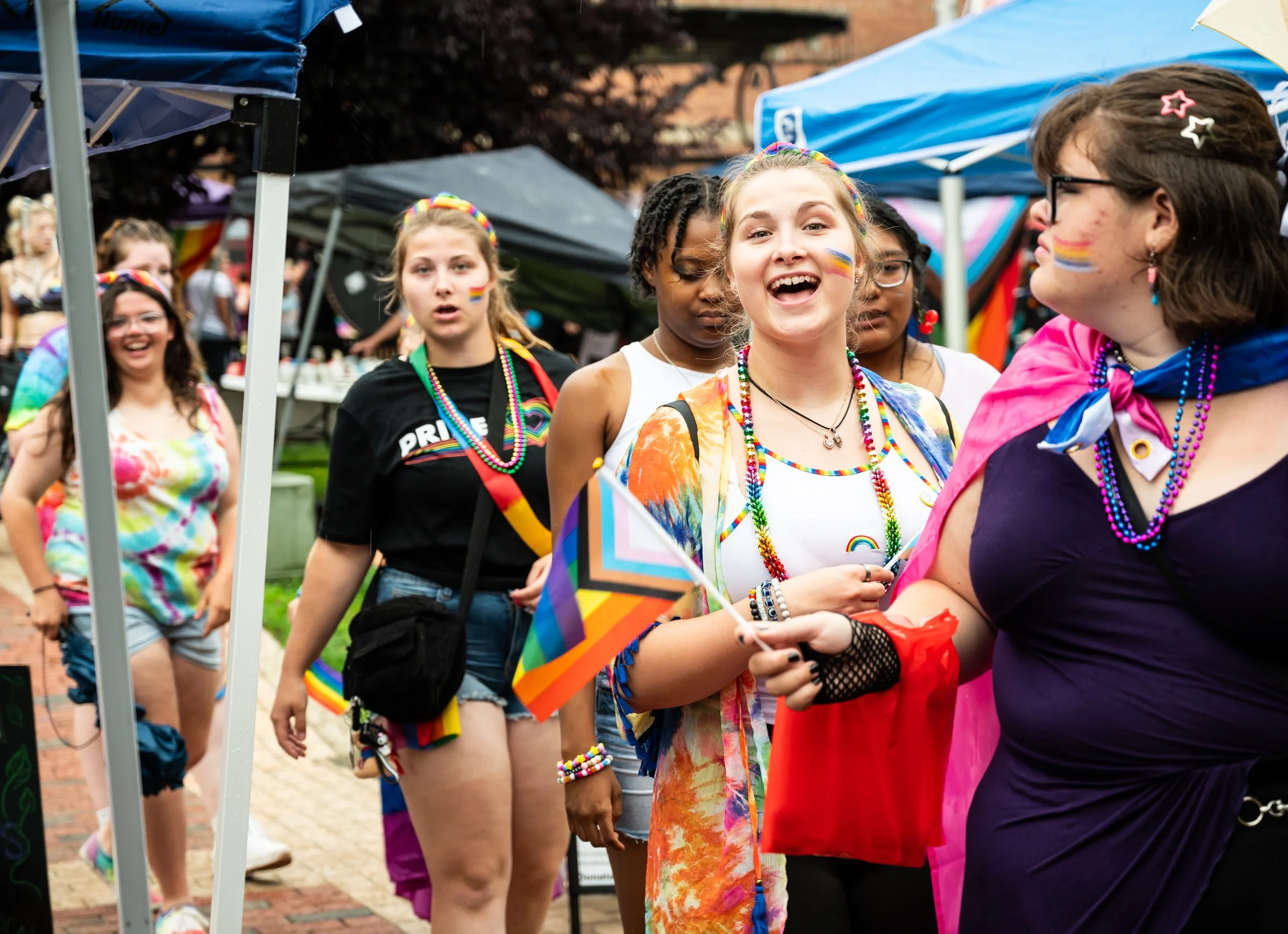 Group of people at a pride parade dressed in rainbow-colored accessories, some with rainbow face paint, holding pride flags, engaging and smiling.