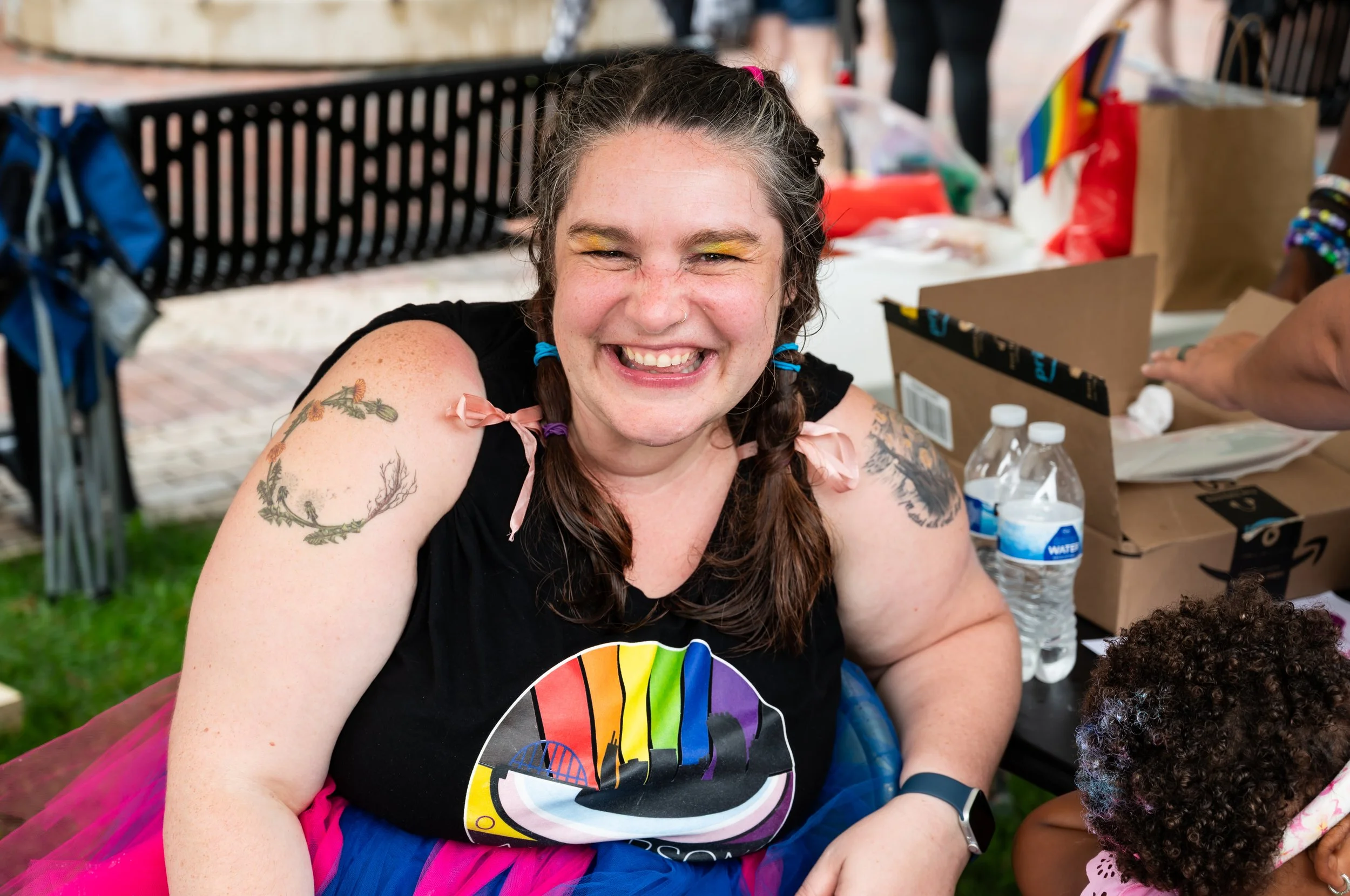Woman with tattoos smiling and wearing rainbow pride T-shirt and rainbow tutu at outdoor event with rainbow flags and water bottles. Anderson Indiana Pride. 