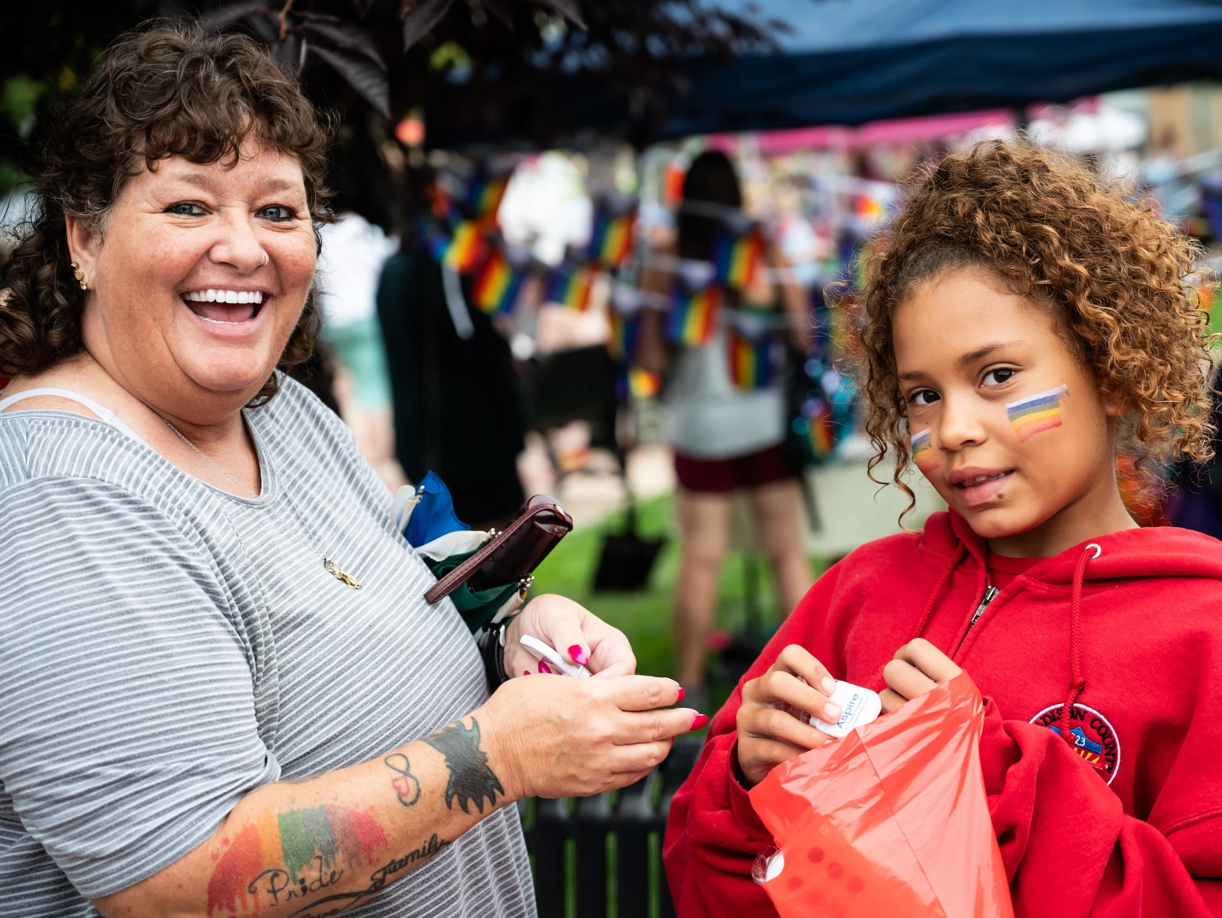 A woman with curly brown hair, tattoos on her arm, and rainbow pride stickers on her face is smiling while handing something to a young girl with curly hair wearing a red hoodie and rainbow pride face paint. They are at an outdoor pride event with ra
