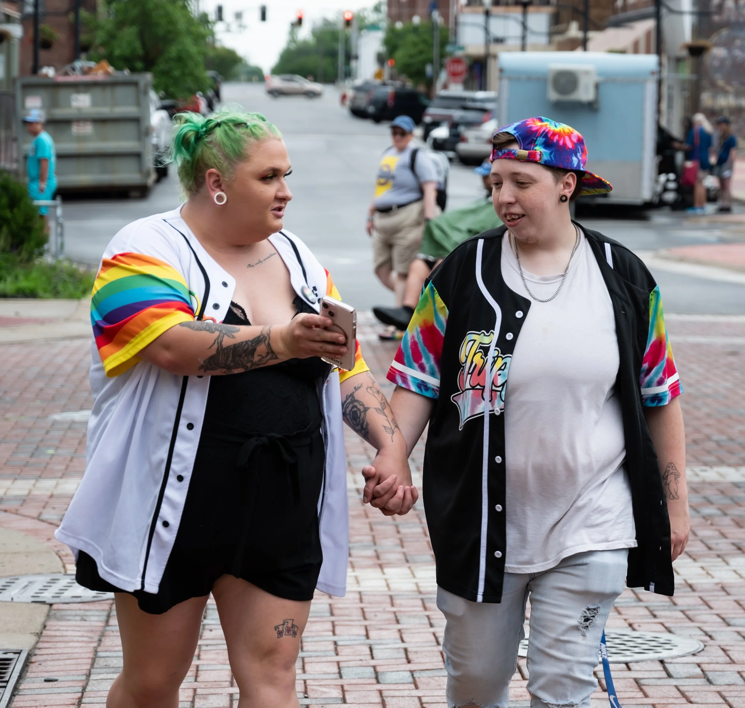 Two people holding hands, walking outdoors on a city sidewalk. One person has green hair, multiple tattoos, and is wearing a rainbow-colored shirt over a black outfit; the other person has a rainbow-colored cap, black and rainbow tie-dye shirt, and d