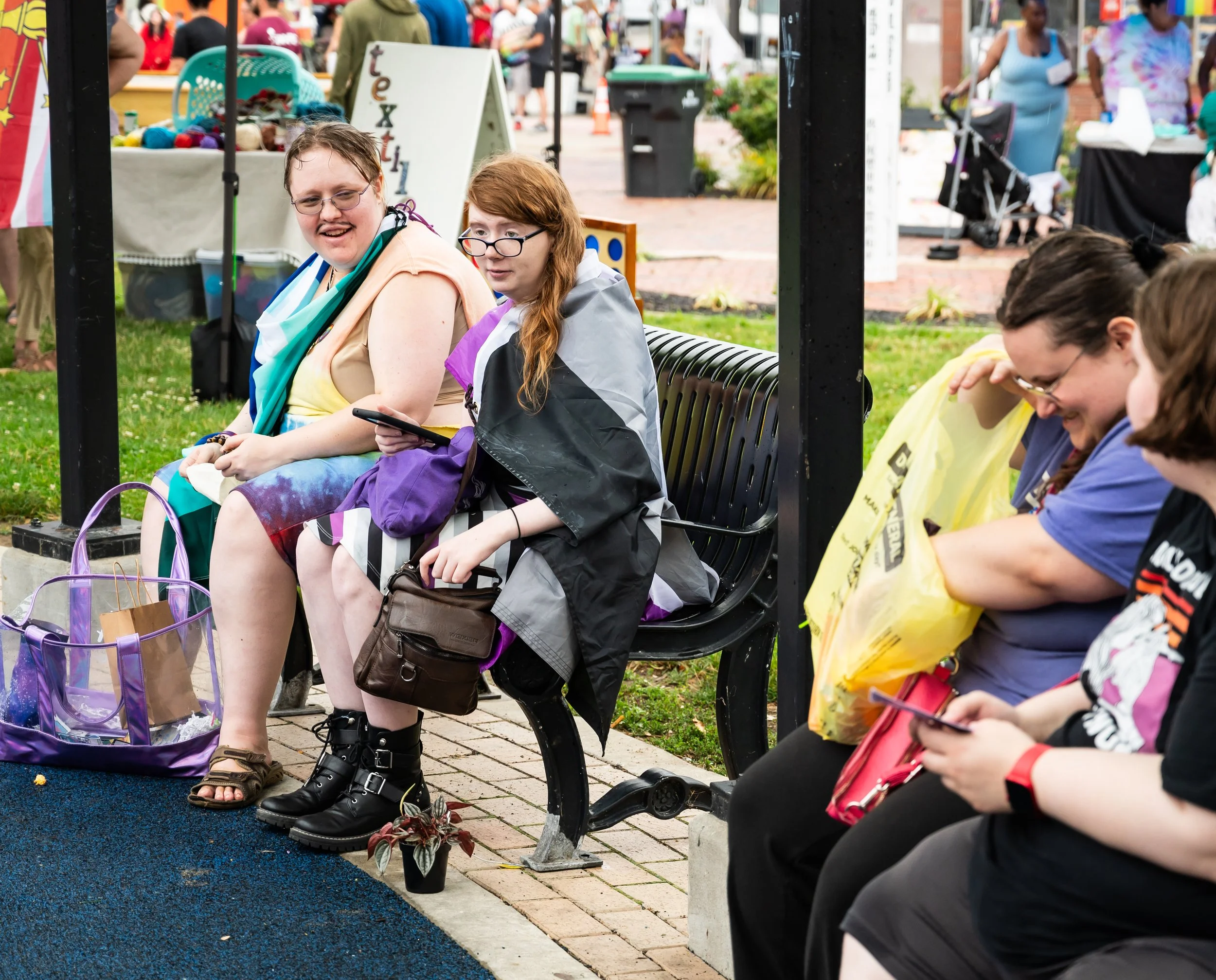 Four women sitting on a bench outdoors, some looking at their phones, with bags and shopping bags nearby, at a busy outdoor event or market.