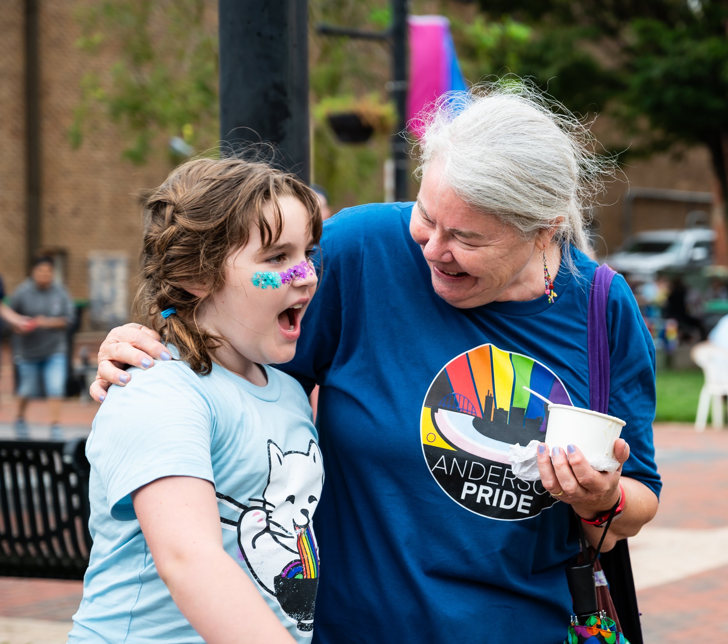 An elderly woman and a young girl sharing a joyful moment at a pride event outdoors. The girl has rainbow face paint and is wearing a T-shirt with a cat and rainbow graphic. The woman has gray hair, wears a blue pride T-shirt, earrings, and holds a c