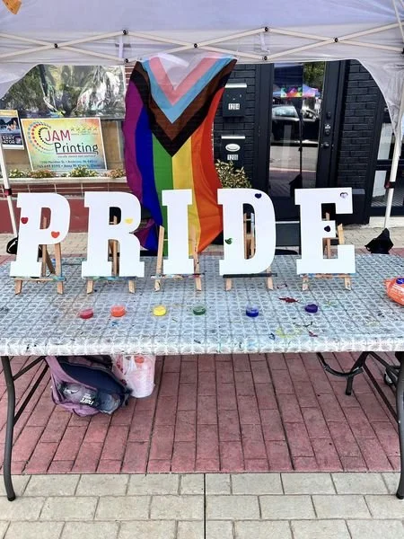 Table with large white letters spelling 'PRIDE' and a rainbow flag in the background, celebrating LGBTQ+ pride at an outdoor event.