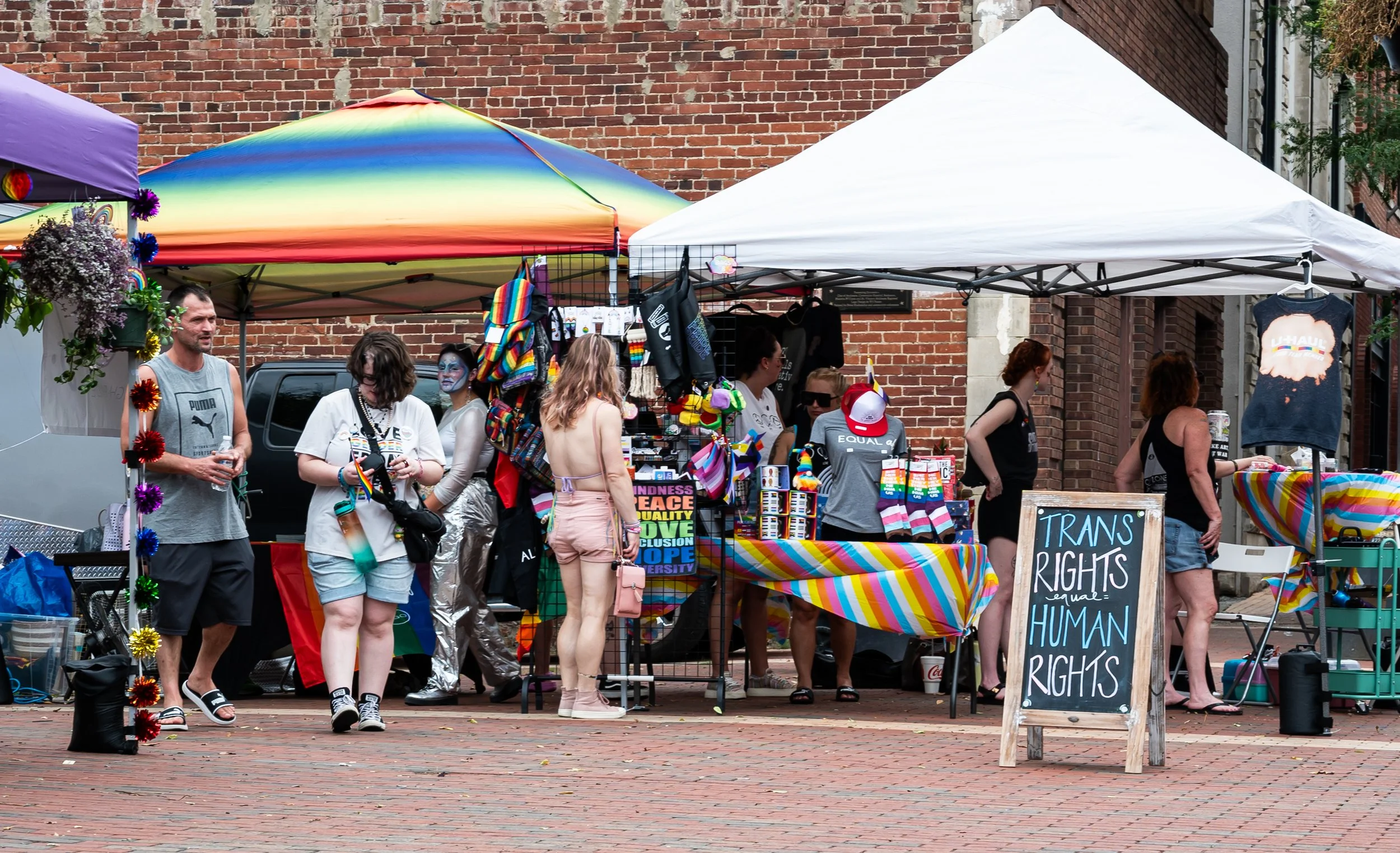 A street market stall with rainbow-colored decorations and LGBTQ+ pride merchandise, including clothing and accessories, with people browsing and a chalkboard sign reading 'Trans Rights Are Human Rights'.