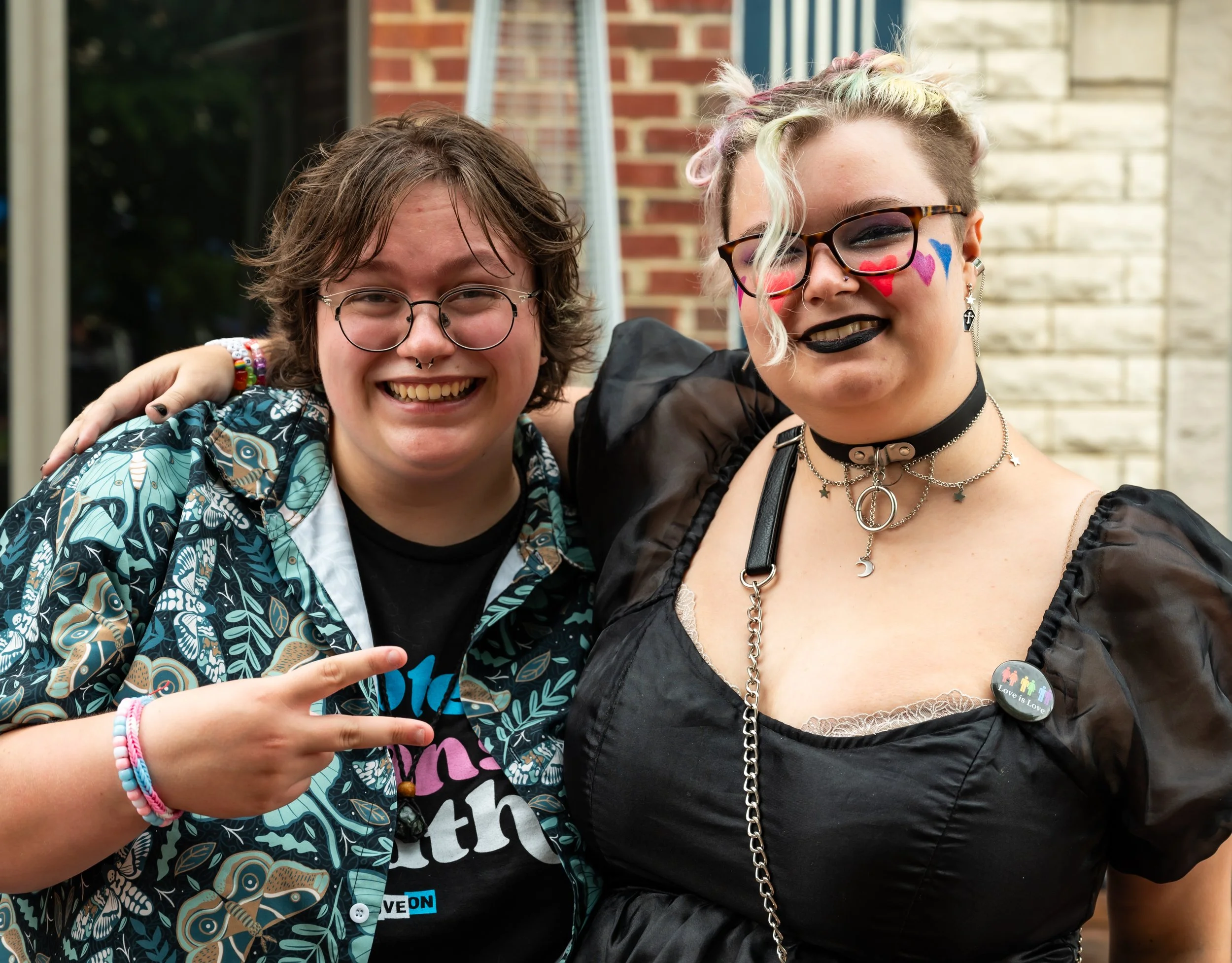 Two smiling people posing outdoors, one with curly hair and glasses making a peace sign, and the other with colorful hair, face paint, and gothic style clothing, standing close together.