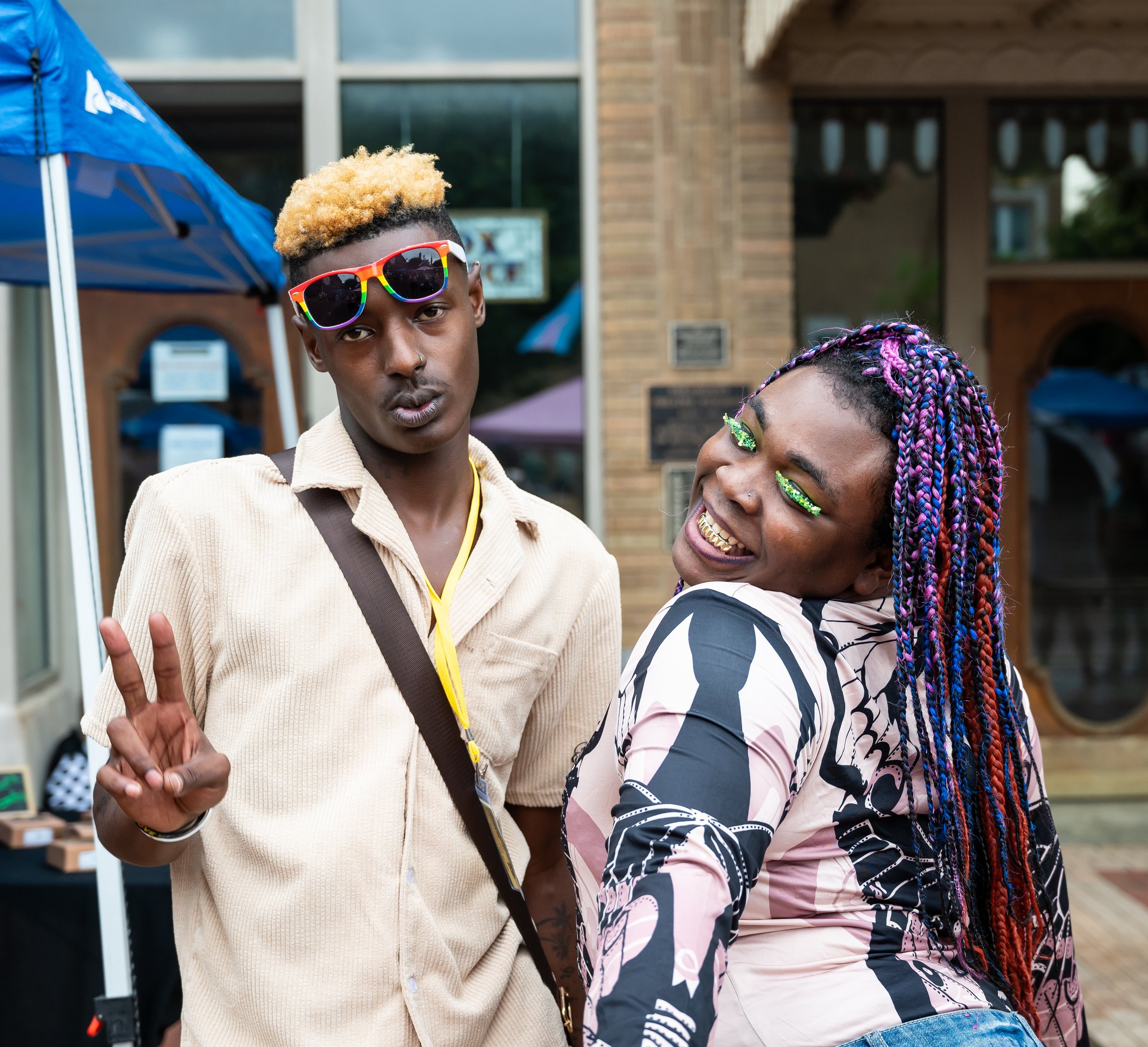 Two young black adults posing outdoors, one making a peace sign and the other smiling with colorful braided hair and bright eye makeup.