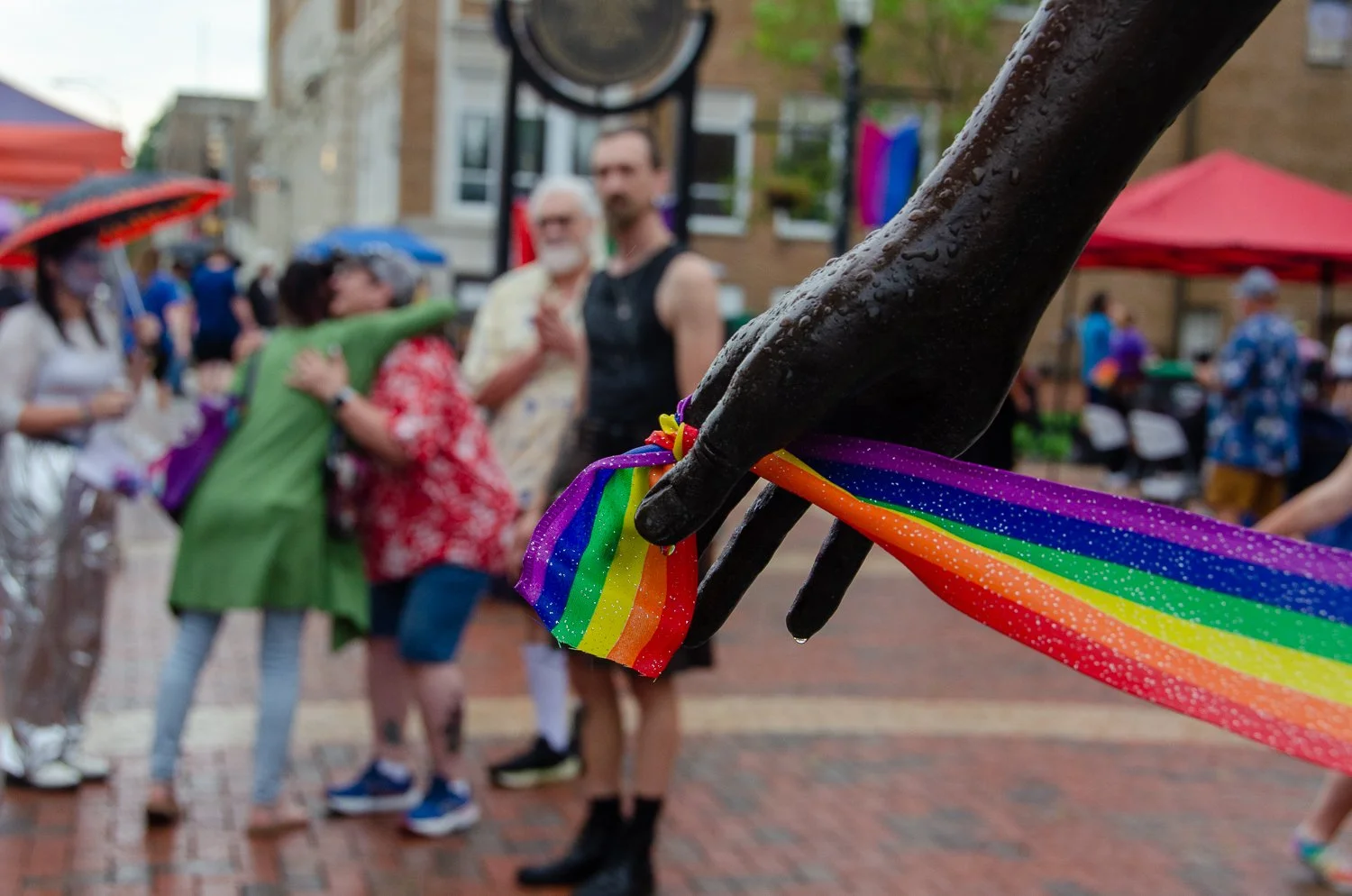 Close-up of a black statue hand holding a rainbow rainbow ribbon at an outdoor gathering with people in the background.
