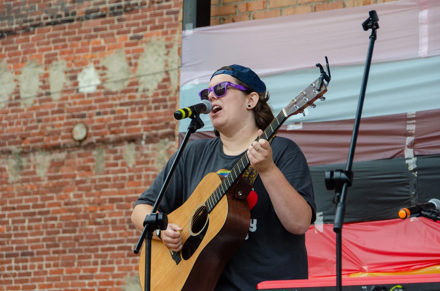 Young woman singing and playing guitar on stage, wearing sunglasses and a backwards cap, in front of a red brick wall, microphone in front of her.