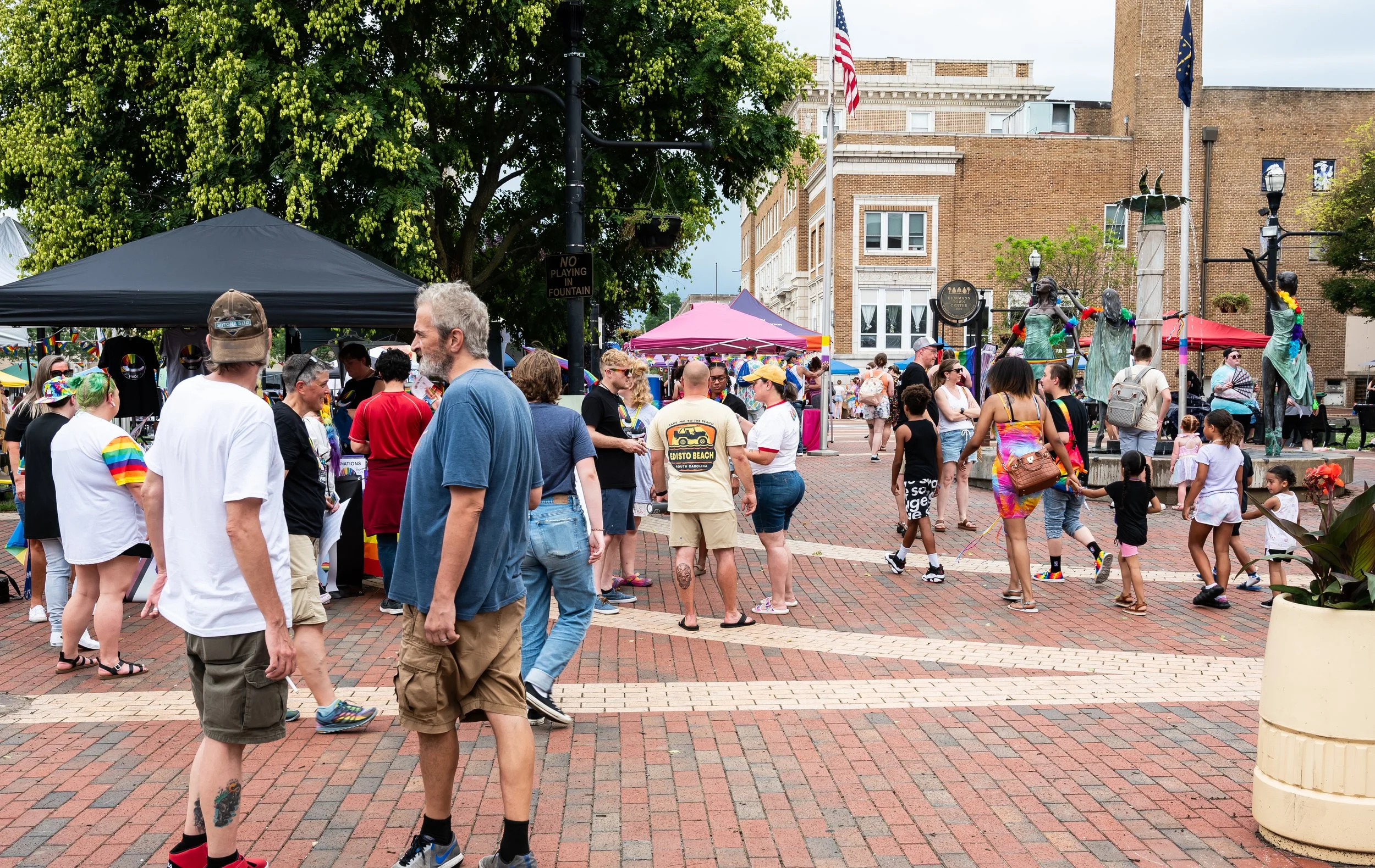 Crowd of people at an outdoor event in a town square, with tents, statues, and buildings in the background.