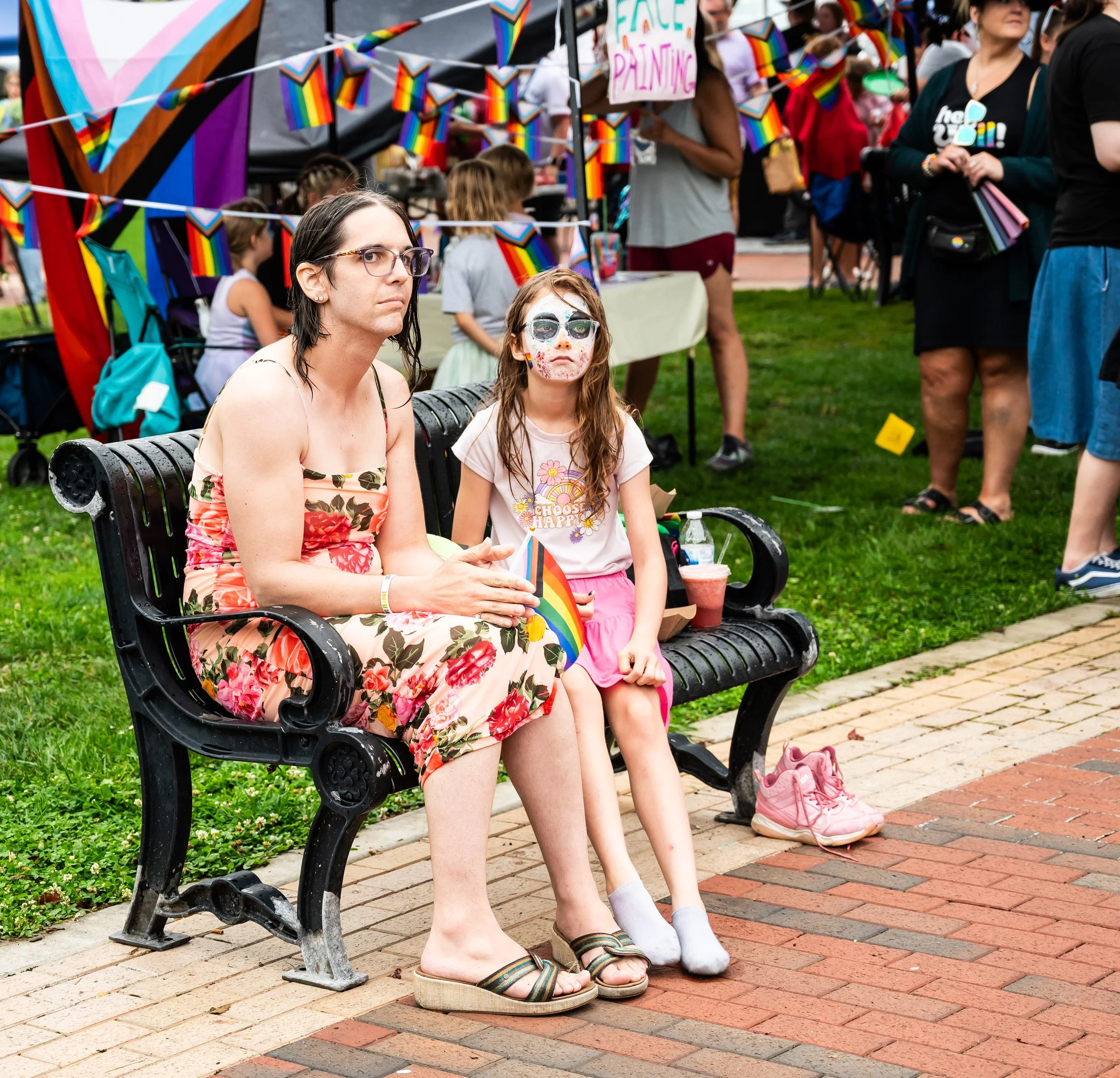 A woman and a young girl sitting on a park bench at a pride parade. The woman wears glasses and a floral dress, and the girl has face paint, a pink skirt, and white socks. Pride flags and rainbow decorations are visible in the background.