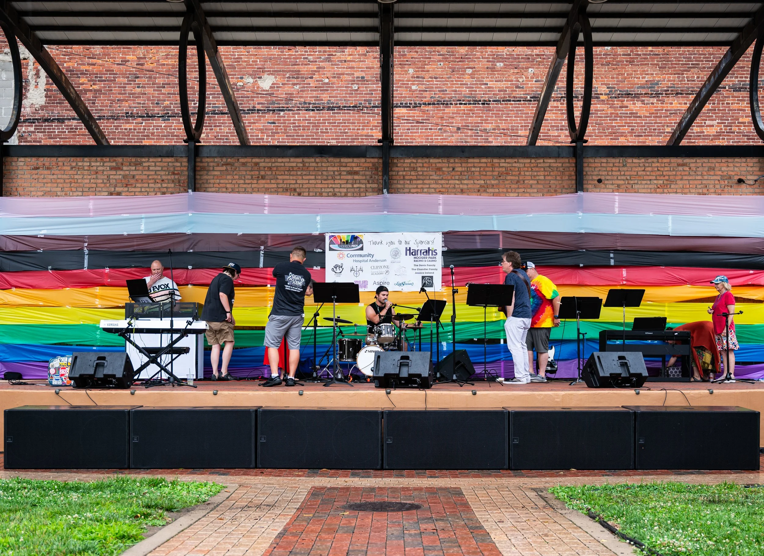 Dickmann Town Center stage decorated with a rainbow flag backdrop, set up outdoors with musicians preparing for a performance, in front of a brick wall and metal framework.
