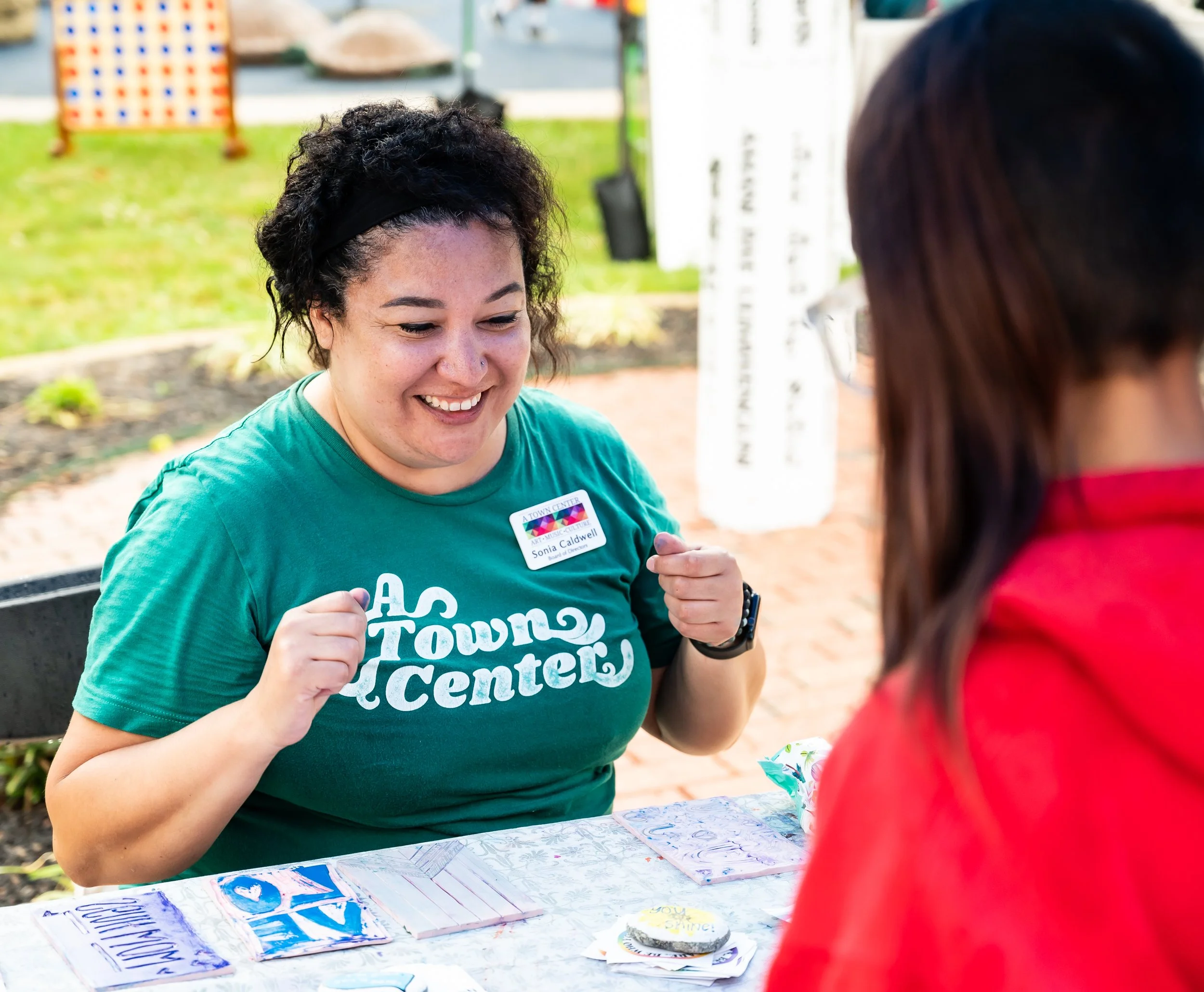 A woman smiling and engaging with another person at an outdoor table for A Town Center, with a colorful sign and a green area in the background.