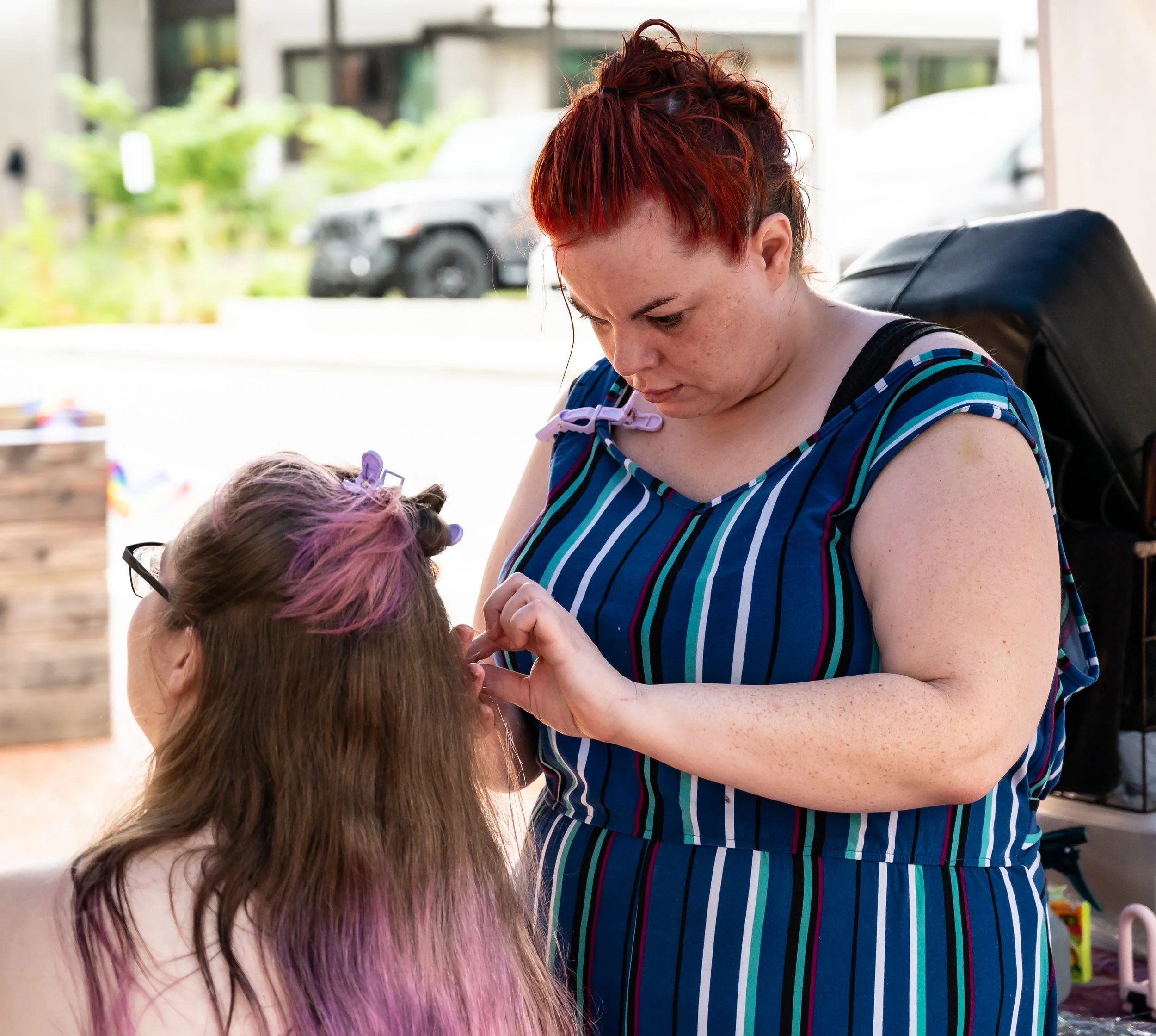 A woman in a striped dress is applying makeup or something similar to a young girl with colorful hair, outdoors near a building and parked cars.