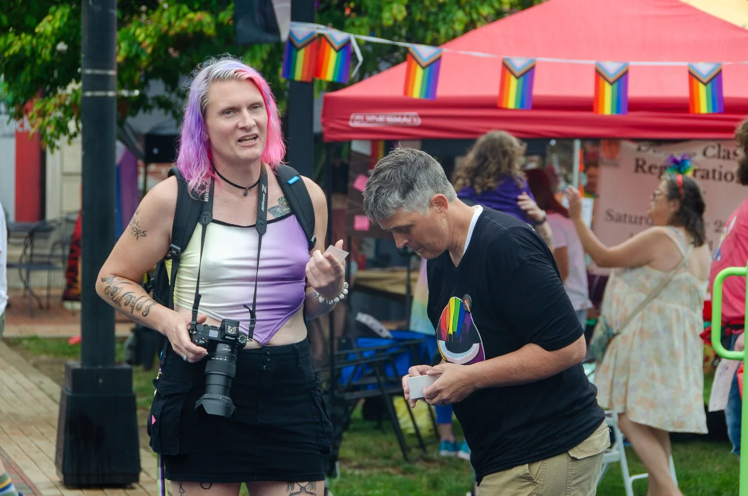 Two people at Anderson Pride, one with pink and purple hair holding a camera, the other with gray hair looking at a small object, with rainbow pride flags in the background.