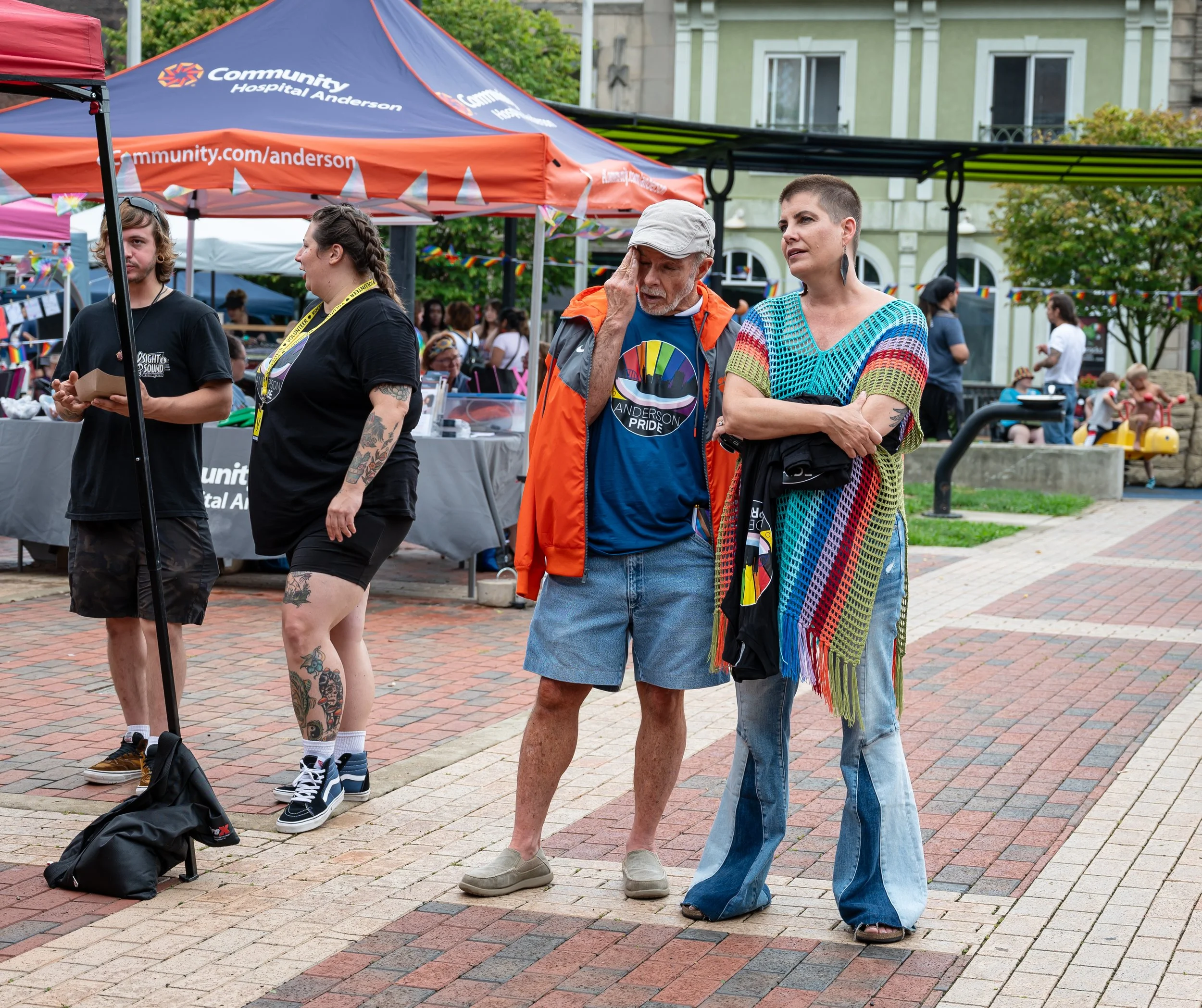 People attending an outdoor event under tents, including a man talking on his phone and a woman wearing a rainbow-colored crochet tunic, with other attendees and booths visible in the background.