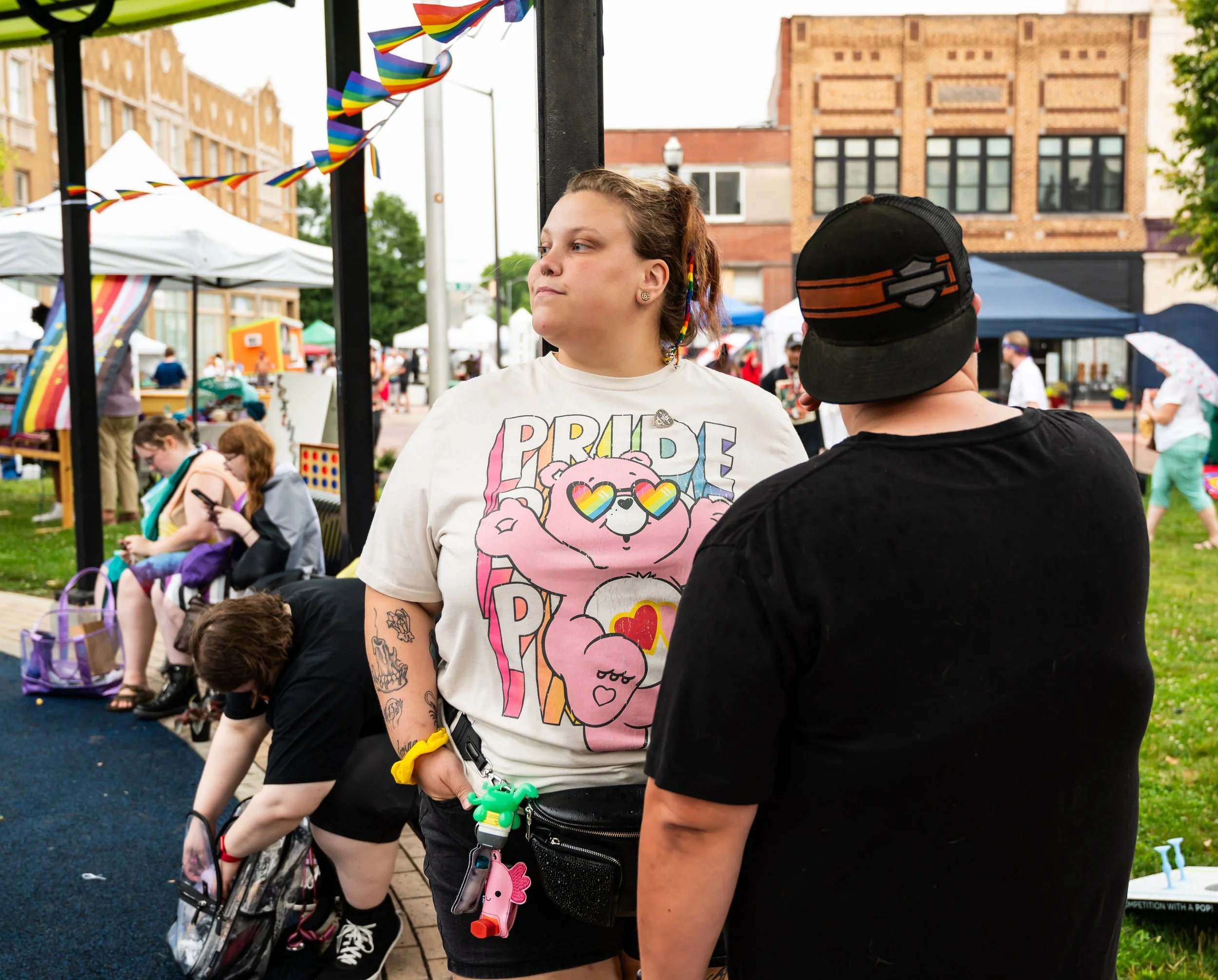 A young woman wearing a Pride-themed t-shirt engaged in conversation at an outdoor Pride event, with tents, rainbow flags, and other attendees in the background.